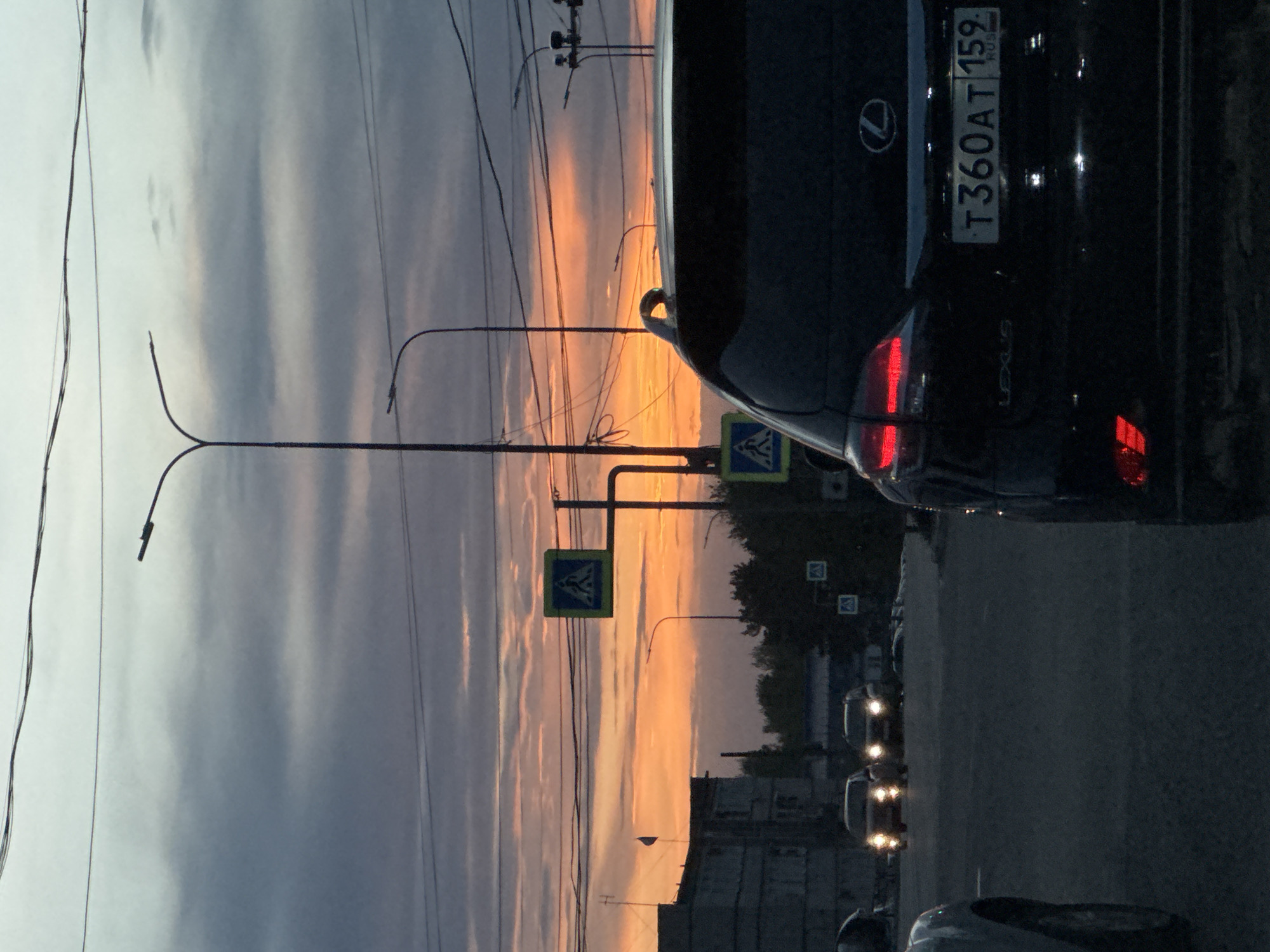 The image depicts a street scene in Chaikovsky, Russia, at twilight. The sky shows a gradient of colors, from grey to orange. A dark-colored Lexus is parked on the right side of the frame, with its license plate "T360AT159" visible. The Lexus logo is also visible on the trunk. Streetlights line the road, with visible wires overhead. There are pedestrian crossing signs. Other cars are visible in the distance. A building is on the left.