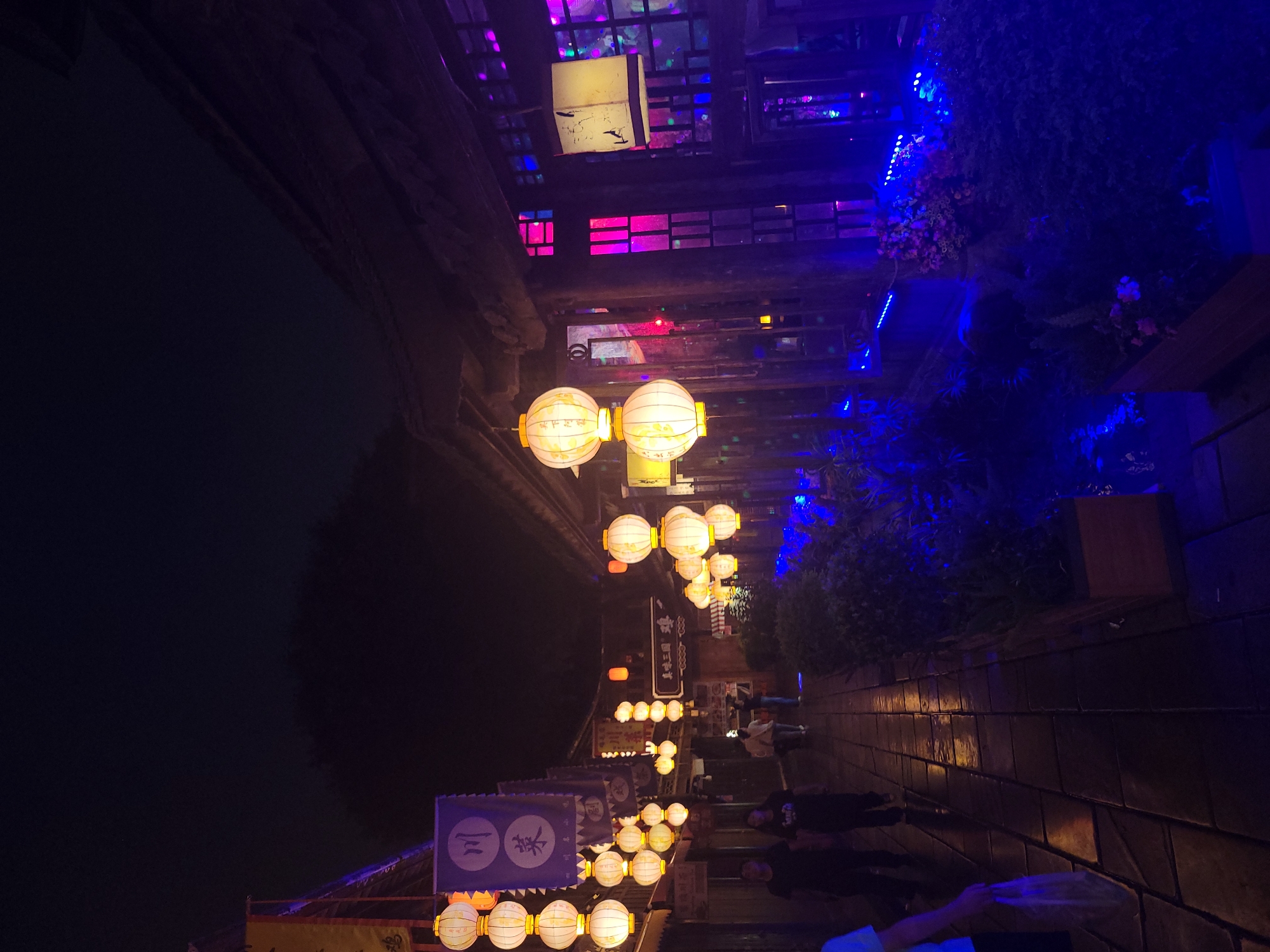 The image depicts a nighttime street scene in Wuhou District, China, illuminated by numerous traditional lanterns and colorful lights. The street is paved with dark, rectangular stones, reflecting the light. On the left side of the street, there are buildings with wooden facades adorned with several strings of white, spherical lanterns. One building prominently displays a blue banner with Chinese characters. Further down the street, more lanterns are visible, along with people walking. On the right side, a building features a series of windows with brightly colored lights, predominantly pink and blue, creating a vibrant and atmospheric glow. The foreground is dominated by a planter box filled with dark green foliage and some flowers, illuminated by bright blue LED lights, which also line the edge of the walkway. The overall impression is of a lively, festive evening street environment.