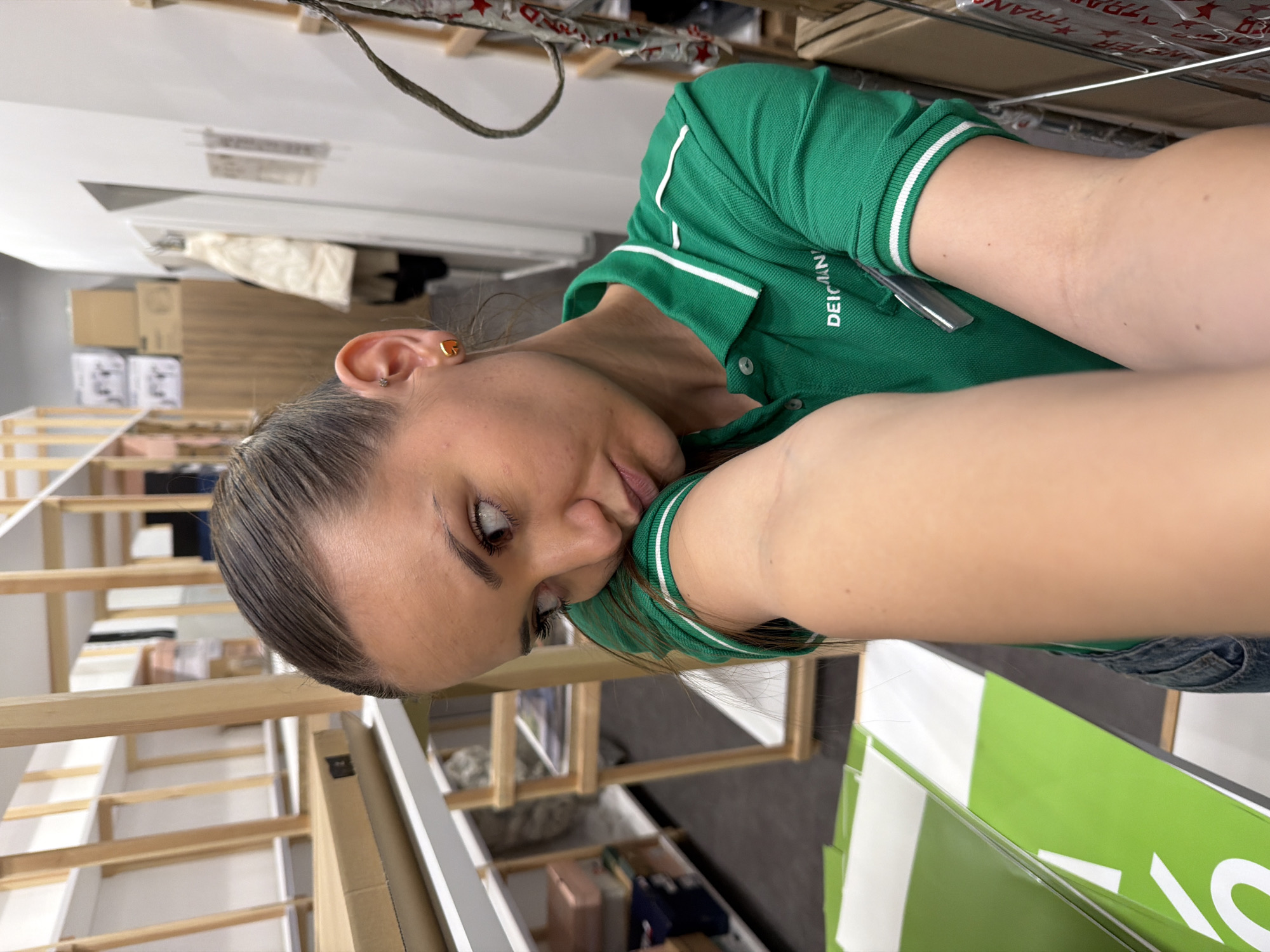 A rotated indoor eye-level selfie shows a woman with brown eyes, a light complexion, and greased-back hair. She is wearing a bright green polo shirt with white accents, and the name "DEICHMAN" is embroidered on it. She's wearing gold-colored earrings in both ears. Her face is close to the camera, and her right arm is partially visible and extended towards the camera.
The background is complex and cluttered. A wooden structure with white elements is visible, possibly shelving or furniture construction in progress. There are cardboard boxes and packaged items stacked in the upper background, and more furniture and storage items underneath the structure. In the lower right is a green and white object with text partially visible.
The location could be a storage area or warehouse, potentially within a retail establishment. There is no definitive visual information in the provided image to confirm that the location is Sofia, Bulgaria.