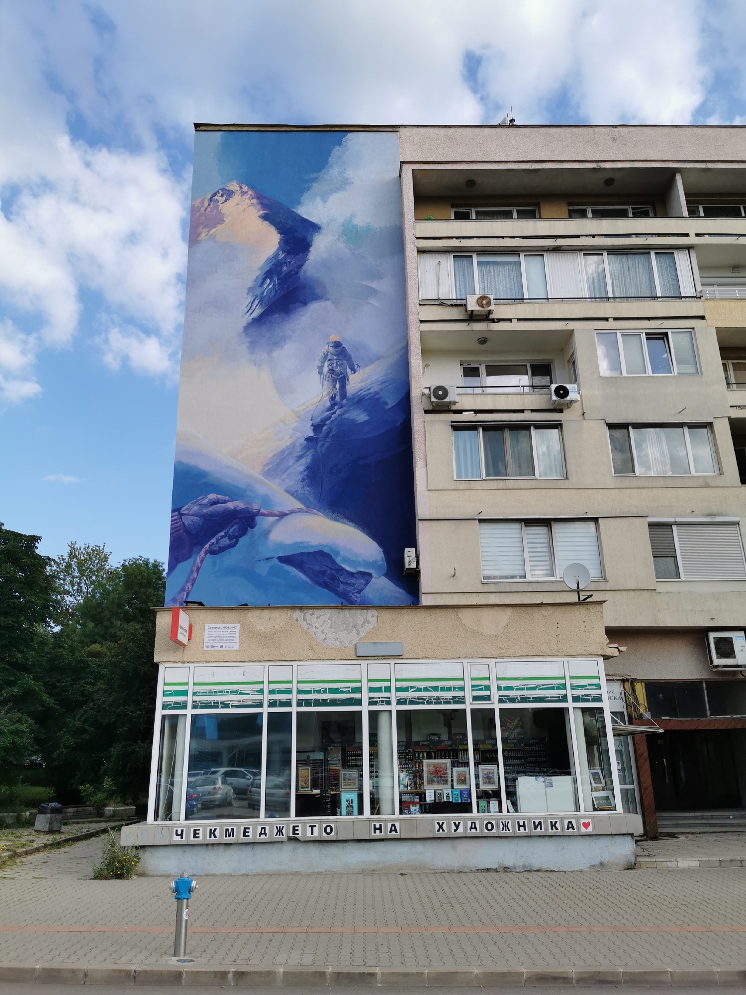 The image depicts a multi-story building with a large mural covering its left side. The mural shows a mountainous landscape with a climber ascending a snowy peak, and a gloved hand holding a rope in the foreground. The building has a light-colored facade with multiple windows and balconies. On the ground floor, there is a shop with large display windows and signage that reads "ЧЕКМЕДЖЕТО НА ХУДОЖНИКА" in Cyrillic script, translating to "The Artist's Chest." The shop appears to sell art supplies. The surrounding environment includes a paved sidewalk, trees, and a blue fire hydrant. The sky is blue with scattered clouds.