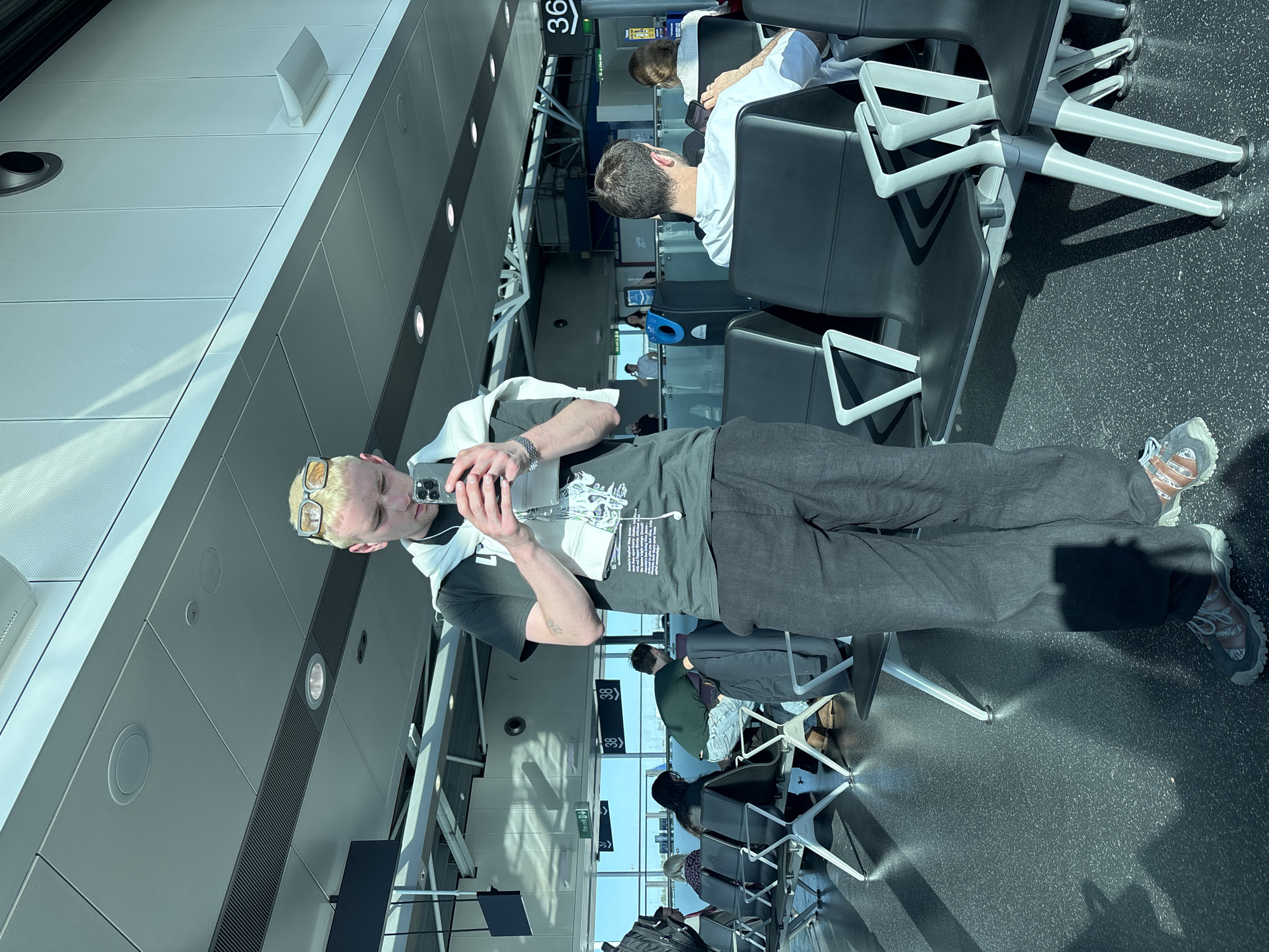 The original image, rotated 90 degrees counter-clockwise, captures a brightly lit airport waiting area, likely a gate. The primary subject is a young person with bleached blonde hair and yellow-tinted glasses, wearing a black and white short-sleeved top with graphic text, dark trousers, and grey sneakers. They are standing and holding a silver smartphone up, appearing to take a photograph or video, possibly a selfie or of the surroundings.
The setting is characterized by modern airport architecture, with white and grey paneled walls and ceiling, featuring recessed lighting fixtures and vents. Rows of black airport chairs with light grey frames fill the space, some of which are occupied by other passengers waiting. Several individuals are visible seated in the background, mostly seen from behind or partially, with some also looking at their phones.
Bright sunlight streams through large windows, casting prominent shadows of the seating on the dark, speckled floor. Visible details include gate numbers "36" and "38" on signs in the background. The scene suggests a daytime setting, likely sunny, given the strong light and shadows. The overall atmosphere is one of waiting and travel in a contemporary airport terminal, possibly in Uttlesford, United Kingdom.