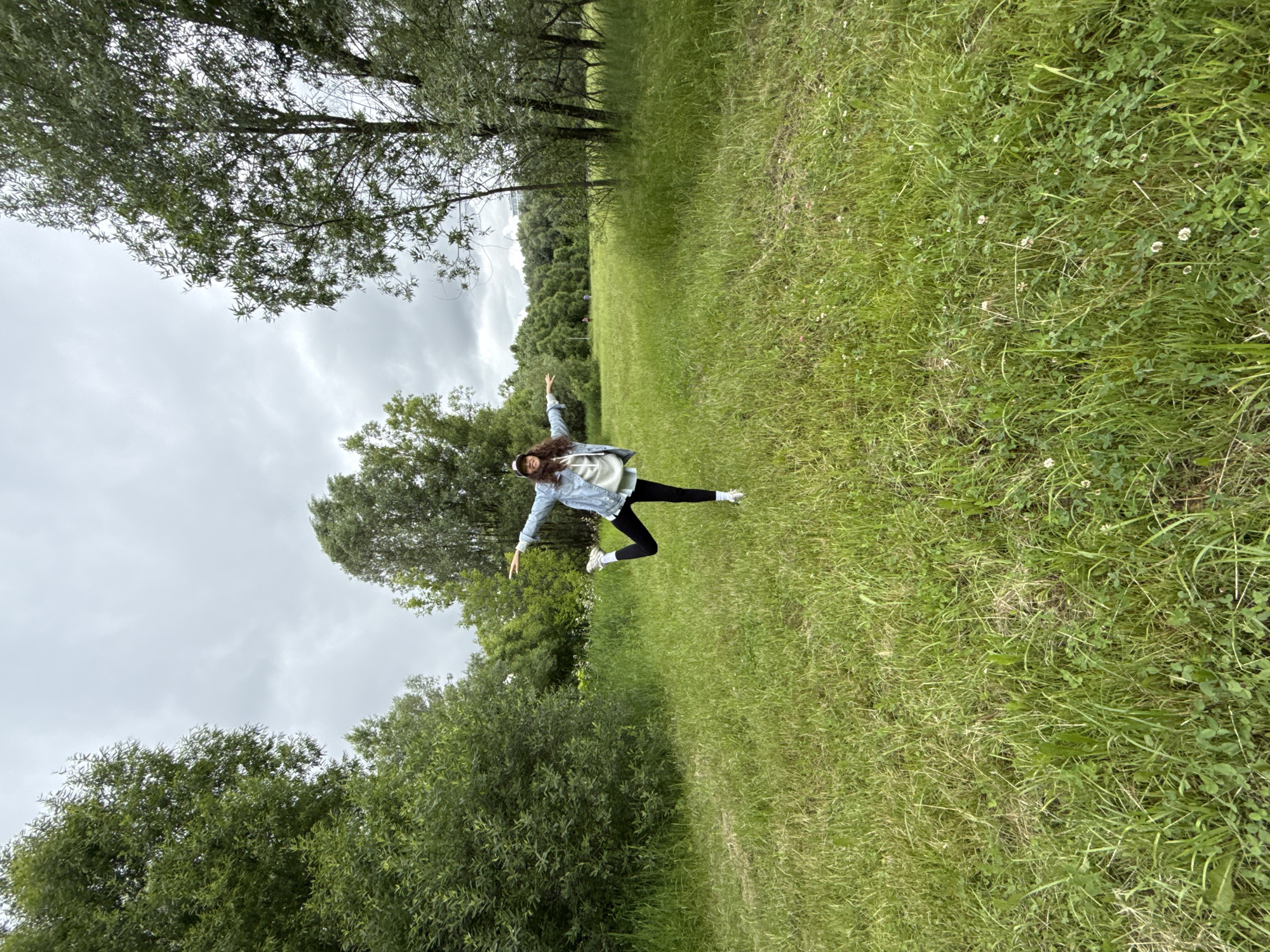 The image shows a young woman with long, curly brown hair, wearing a denim jacket over a light-colored hoodie, black leggings, and white sneakers. She is captured mid-air, in a dynamic pose with her arms outstretched and legs apart, as if jumping or dancing. The photo is taken outdoors in a grassy field with trees in the background. The sky is overcast, suggesting a cloudy day. The lighting is soft, indicating it might be daytime, possibly late afternoon or early morning. The setting appears to be a park or a large open area with lush green grass and scattered small white wildflowers. The woman's expression is not clearly visible due to the distance and pose, but her energetic posture suggests joy and freedom. The location is hinted at being Moscow, Russia, though there are no explicit textual or landmark cues to confirm this. The overall mood is lively and carefree, set against a natural, somewhat subdued backdrop due to the weather.