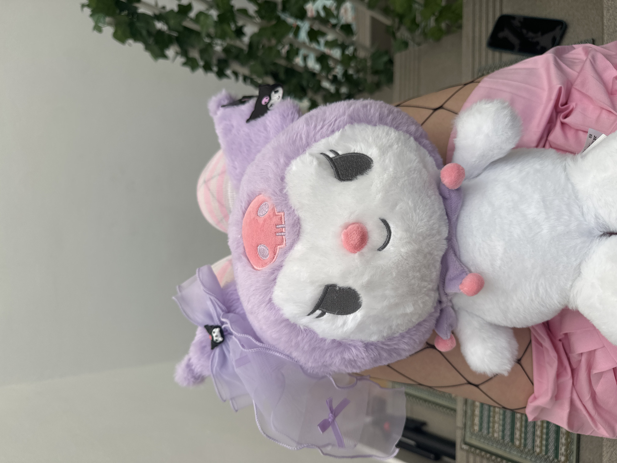 A close-up shot features a purple Kuromi plush toy with white facial features, resting on what appears to be a leg covered in fishnet stockings and a pink garment. The leg is possibly positioned indoors, given the presence of interior elements in the background, such as a wall and foliage. The exact location in Zagreb, Croatia, cannot be determined from the provided image.