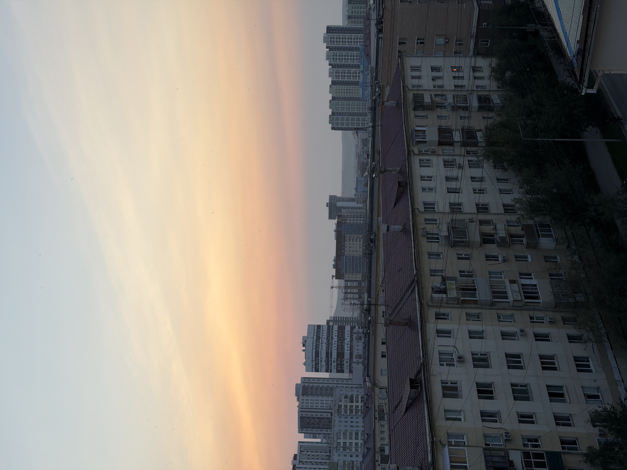 The image depicts a cityscape at dusk, likely in Yakutsk, Russia, given its characteristic architectural style and the pastel hues of the sky, indicative of high-latitude sunsets. The foreground is dominated by the roofline of a low-rise apartment building with a red tiled roof. Behind it, a larger, beige apartment building with multiple windows, balconies, and visible external wiring extends across the frame. Further into the distance, several taller, modern apartment buildings are visible, some with a lighter color palette and grid-like window patterns. Construction cranes are present amongst the buildings, suggesting ongoing development. The sky transitions from a soft yellow-orange near the horizon to a pale blue overhead, with a few faint clouds. The overall scene is one of urban residential architecture under a tranquil twilight sky. No people are visible.