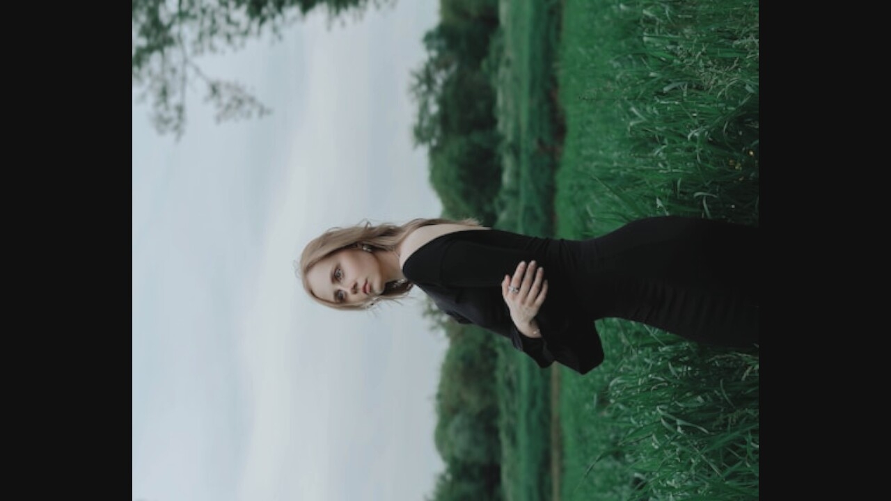 A young woman with blonde hair stands in a field of tall green grass, wearing a black dress. She is facing left, looking out into the distance. Her left arm is bent, with her hand resting on her chest, and her right arm is down by her side. The background consists of trees and a cloudy sky.