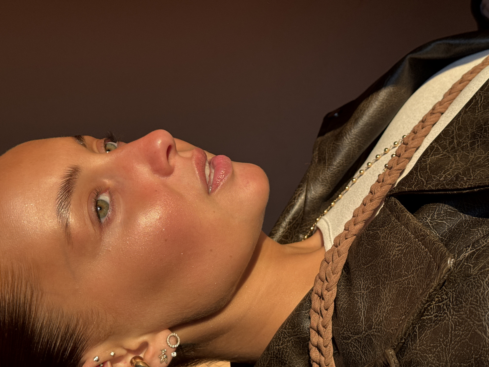 This image is a close-up, profile portrait of a person, likely female, captured from the chest up with the head tilted slightly upwards and to the left (from the viewer's perspective). Strong, warm, directional lighting illuminates the subject, creating bright highlights on the cheekbones, nose, and forehead, giving the skin a dewy or glistening appearance. The person has light-colored eyes, possibly green or hazel, and well-groomed eyebrows. Her lips are slightly parted, revealing a subtle pink hue and shiny finish.
Multiple earrings are visible on her ear: a prominent golden hoop or cuff, a circular earring with small stones, and several smaller stud piercings. Her hair is pulled back, and the hairline along her temple is visible. She is wearing a dark brown, textured jacket, possibly leather or faux-leather, over a light-colored (cream or white) top. A braided brown strap and a thin gold beaded chain are draped near her shoulder, suggesting accessories.
The background is a plain, solid dark brown, providing a minimalist setting that keeps the focus entirely on the subject. The overall impression is one of calm and subtle expressiveness, emphasizing skin texture and the interplay of light. No specific activity is depicted beyond posing for the photograph. There is no visible text within the image. This image is associated with Chaikovsky, Russia.