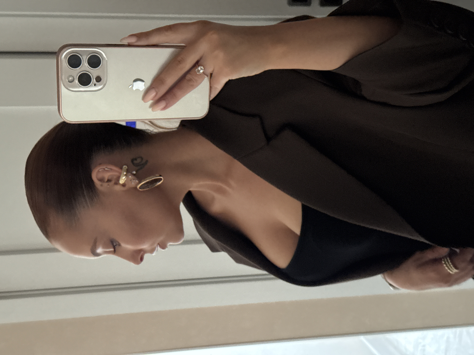 The image shows a woman with her hair pulled back, wearing a dark brown blazer over a black top. She is wearing large gold earrings and multiple rings on her fingers. She is holding a smartphone, which is facing the camera, in her right hand. The background appears to be indoors, possibly in a home or hotel room, with a white door or wall visible. The image does not contain any information about the city of Chaikovsky, Russia.