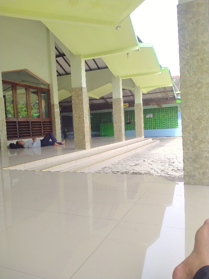 The image shows a covered outdoor area of a building located in Ngrampal, Indonesia, at latitude -7.409 and longitude 111.053. The area is tiled with a light-colored, smooth, polished surface, partially wet from a recent rain. A man lies on the floor to the left of the image, with his head on the floor and his legs extended towards the right. He is wearing dark pants and a light-colored shirt. The roof of the building is supported by multiple columns, creating a covered space. The roof is made of a white material and has a green underside. The walls of the building are also tiled with a light-colored material. A second person, only their right foot and lower leg visible, is sitting in the foreground of the image.