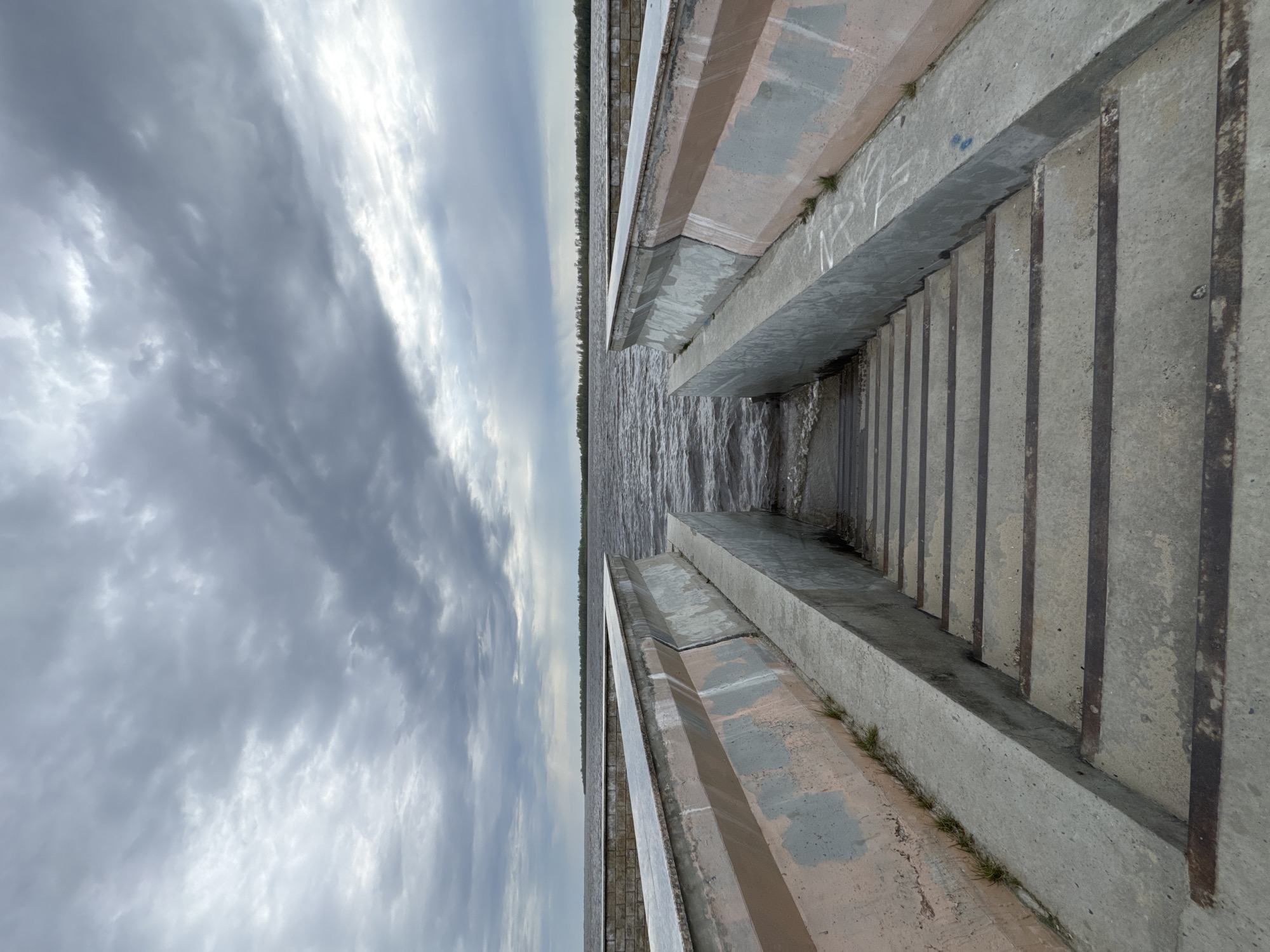 The image shows a concrete structure with vertical metal bars, possibly part of a pier or embankment, located in Nizhnevartovsk, Russia. The structure leads down to the water, which reflects the cloudy sky. The sky is predominantly gray with white patches.