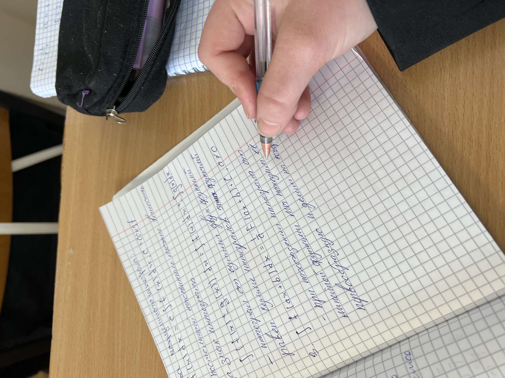 A person is writing in a graph-paper notebook with a clear-barreled ballpoint pen. The notebook is open on a wooden desk surface. A black pencil case sits beside the notebook. The notebook appears to contain handwritten mathematical or scientific notes in the Russian language. The location may be inferred as Chaikovsky, Russia based on image metadata.