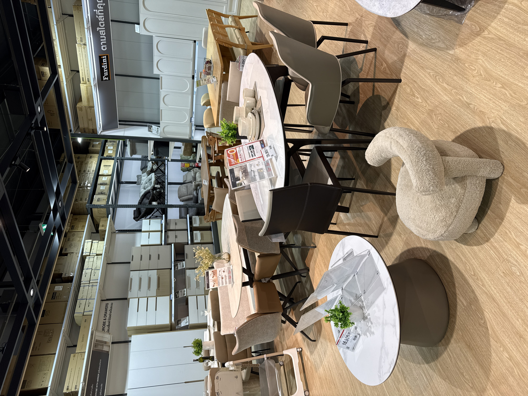 The image depicts an interior view of a furniture store, likely in Baan Chaweng Noi, Thailand. The store is well-lit and organized, with various furniture sets displayed. In the foreground, a round white marble-top coffee table with a textured gray base is prominent. Next to it, a cream-colored armchair with a unique curved back is visible. Behind the coffee table, a dining set features an oval marble-top dining table surrounded by upholstered chairs. Further back, other dining sets and seating arrangements are displayed, including a large wooden dining table with benches. Shelving units filled with boxes and storage containers line the walls. A sign with "Furdini" and Thai text is visible, indicating a brand or store name. The flooring is light-colored wood laminate. No people are present in the image.