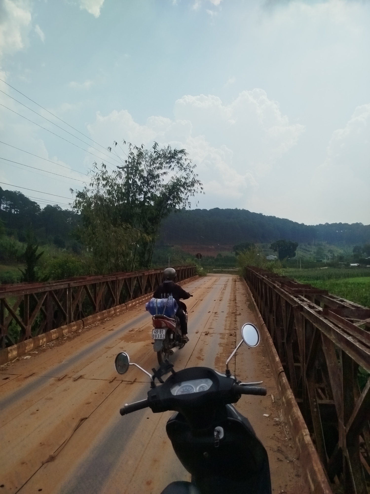 This image captures a first-person perspective from a motorcycle crossing a rusty, metal truss bridge in a rural, hilly landscape. Ahead, another motorcycle, ridden by a person wearing a helmet, navigates the dirt and metal-plated bridge surface. This leading motorcycle is carrying a blue tarp or bag secured on its rear, and its Vietnamese license plate, "49-B1 093.02," is clearly visible.
The scene is set in Thôn 1, Vietnam, under a bright, partly cloudy daytime sky. To the left of the bridge, power lines are visible, extending towards distant, forested hills. A prominent cluster of bamboo trees stands by the bridge on the left side. To the right, green agricultural fields stretch out, leading to more distant hills dotted with trees and a few scattered structures. The bridge itself is an aged, industrial-looking structure with heavily rusted metal beams, suggesting it has been in use for a considerable time. The overall impression is one of travel through a scenic, rural environment.