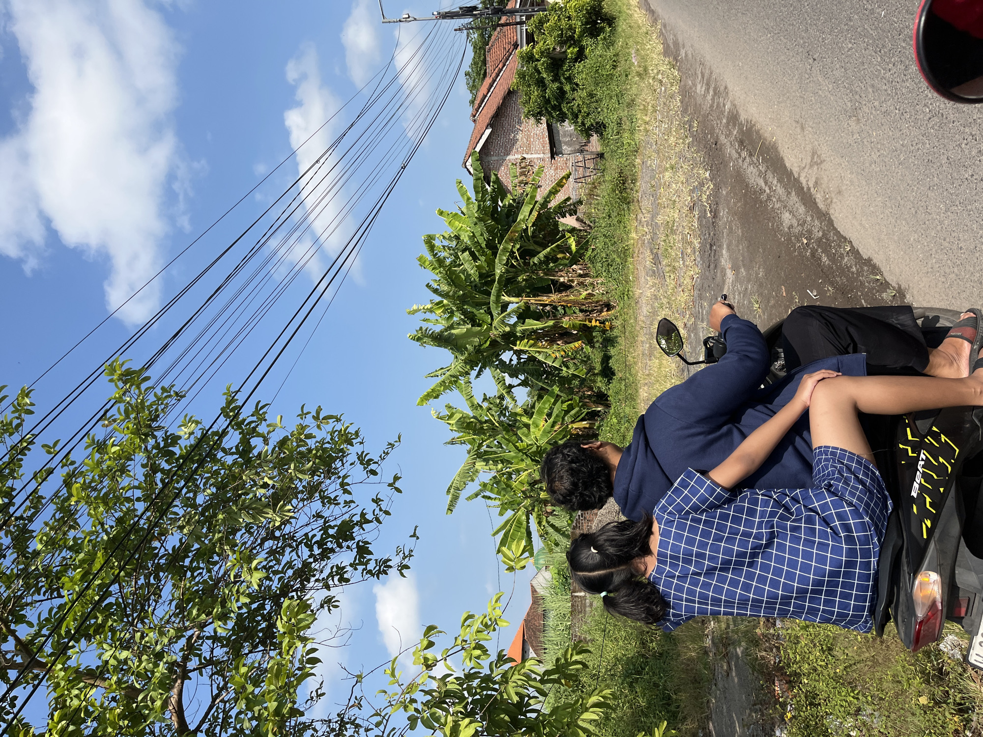In Jati Alun Alun, Indonesia, two people ride a black scooter along a paved road. The person in front, with dark curly hair, wears a navy blue hoodie and black pants. They are driving the scooter. Seated behind them, a child with dark hair in pigtails wears a blue and white checkered dress and is holding onto the driver. The environment is rural, with lush green vegetation including banana trees and other foliage. Several houses with terracotta roofs are visible in the background, along with power lines stretching across the clear blue sky, dotted with fluffy white clouds. The road appears to be a rural street.