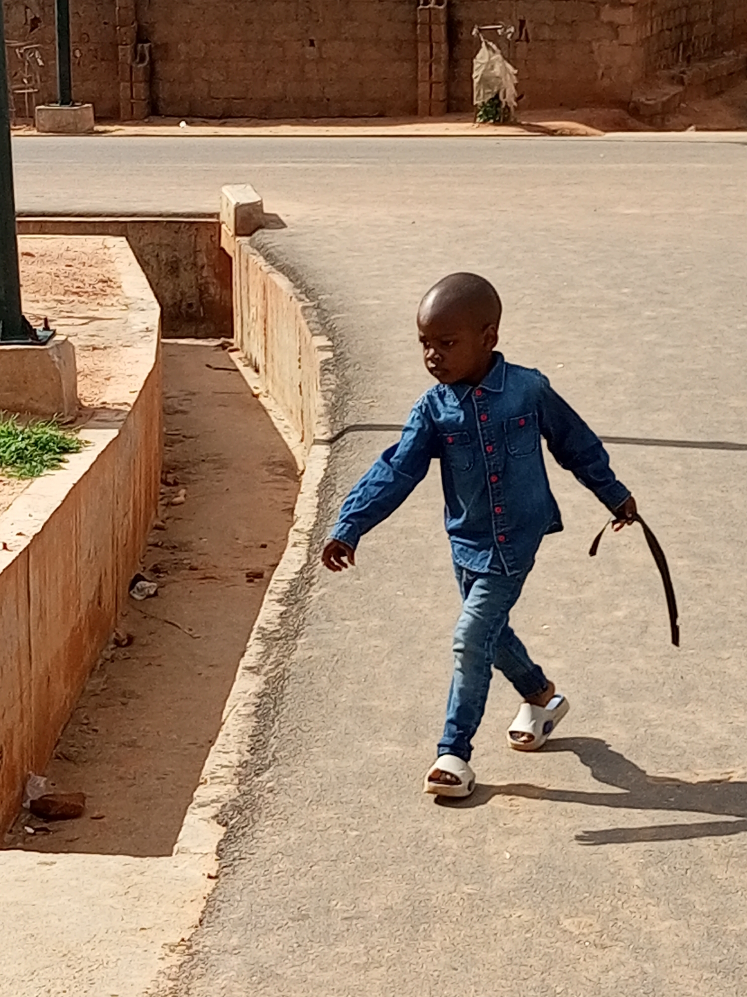 A young boy walks on a paved surface next to a dirt drainage ditch in Tempure, Nigeria. The boy is wearing a denim shirt, denim pants, and white sandals. He is holding a dark strap in his right hand. The drainage ditch is lined with concrete and has some debris in it. In the background, a brick wall and a street are visible. The lighting suggests it is daytime.