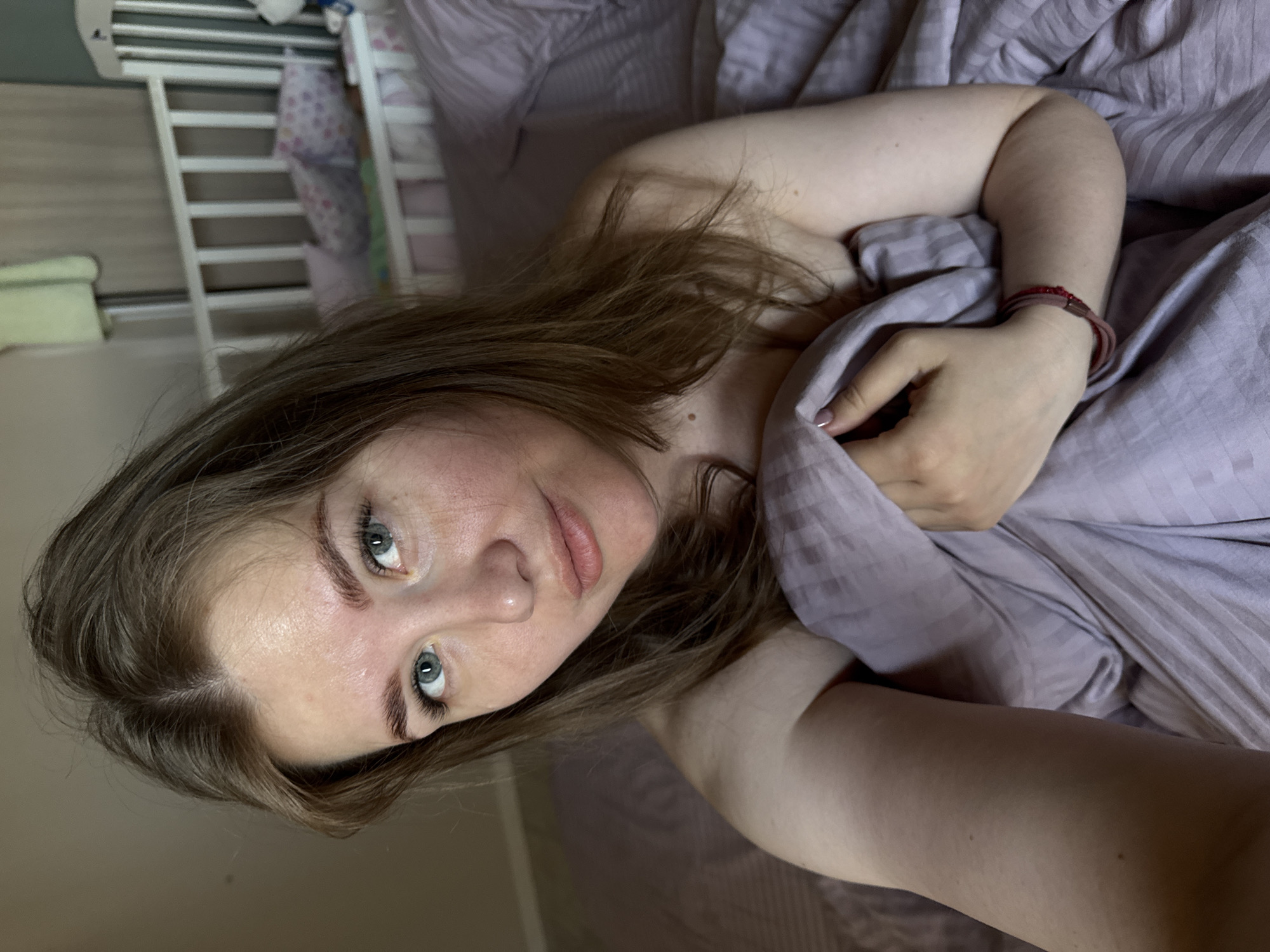 A woman with light skin, brown hair, and blue eyes is lying in bed in a location that may be in Ферма, Russia. She is partially covered by a light purple blanket. Her left arm is visible, and she is wearing a red bracelet on her wrist. A crib with white bars is visible in the background. The room has a neutral tone.