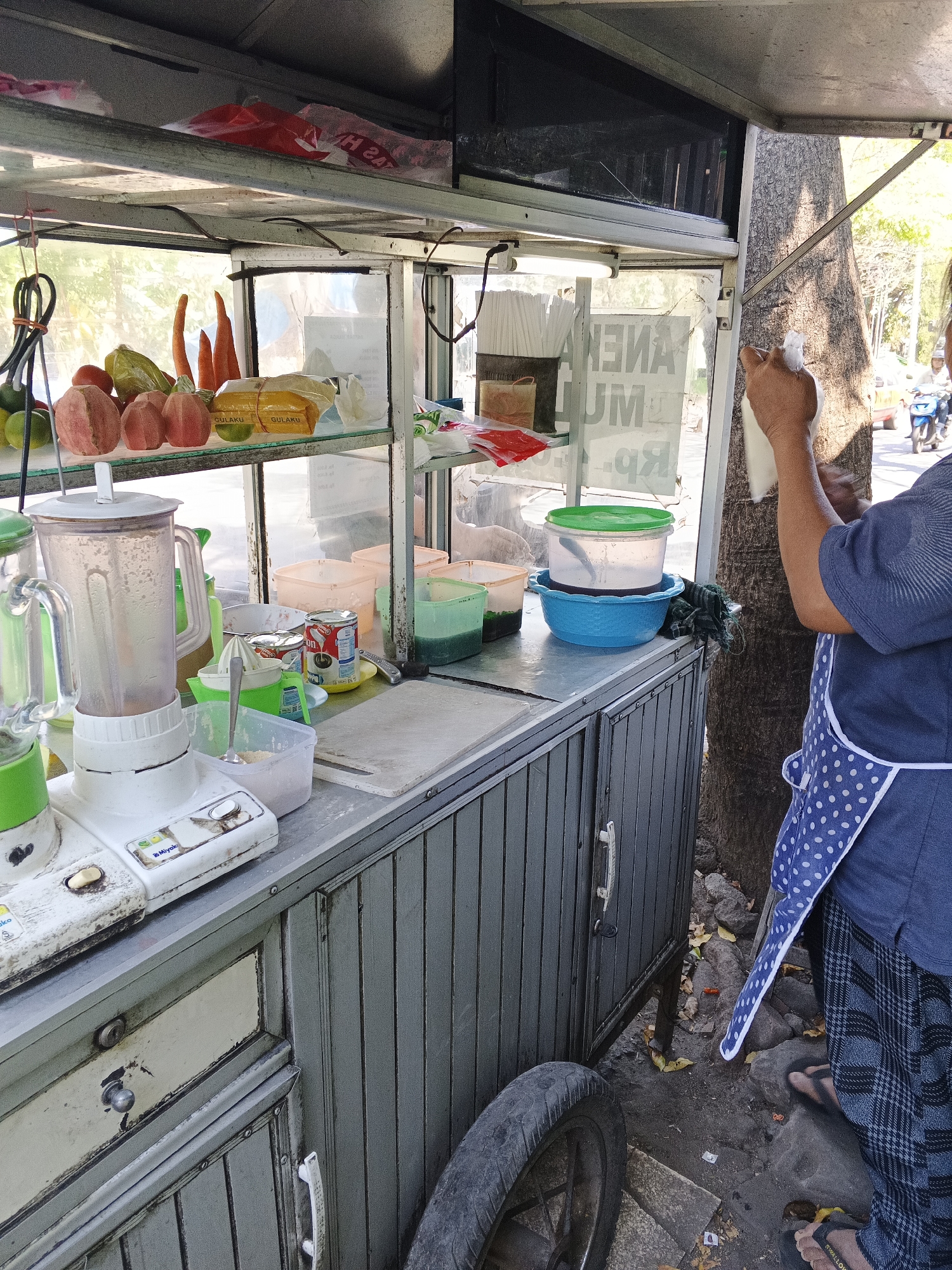 In Tambak Kemerakan, Indonesia, a street vendor operates a mobile cart, likely for selling beverages. The cart is laden with various items, including a blender, containers of liquids, and fresh produce such as red apples and carrots. A person wearing a blue shirt, a white polka-dot apron, and plaid pants is seen preparing something, holding a white object in their hands. The background reveals a street scene with a tree, parked vehicles, and a glimpse of buildings, suggesting an urban environment. The overall atmosphere is that of a typical street market in Indonesia.