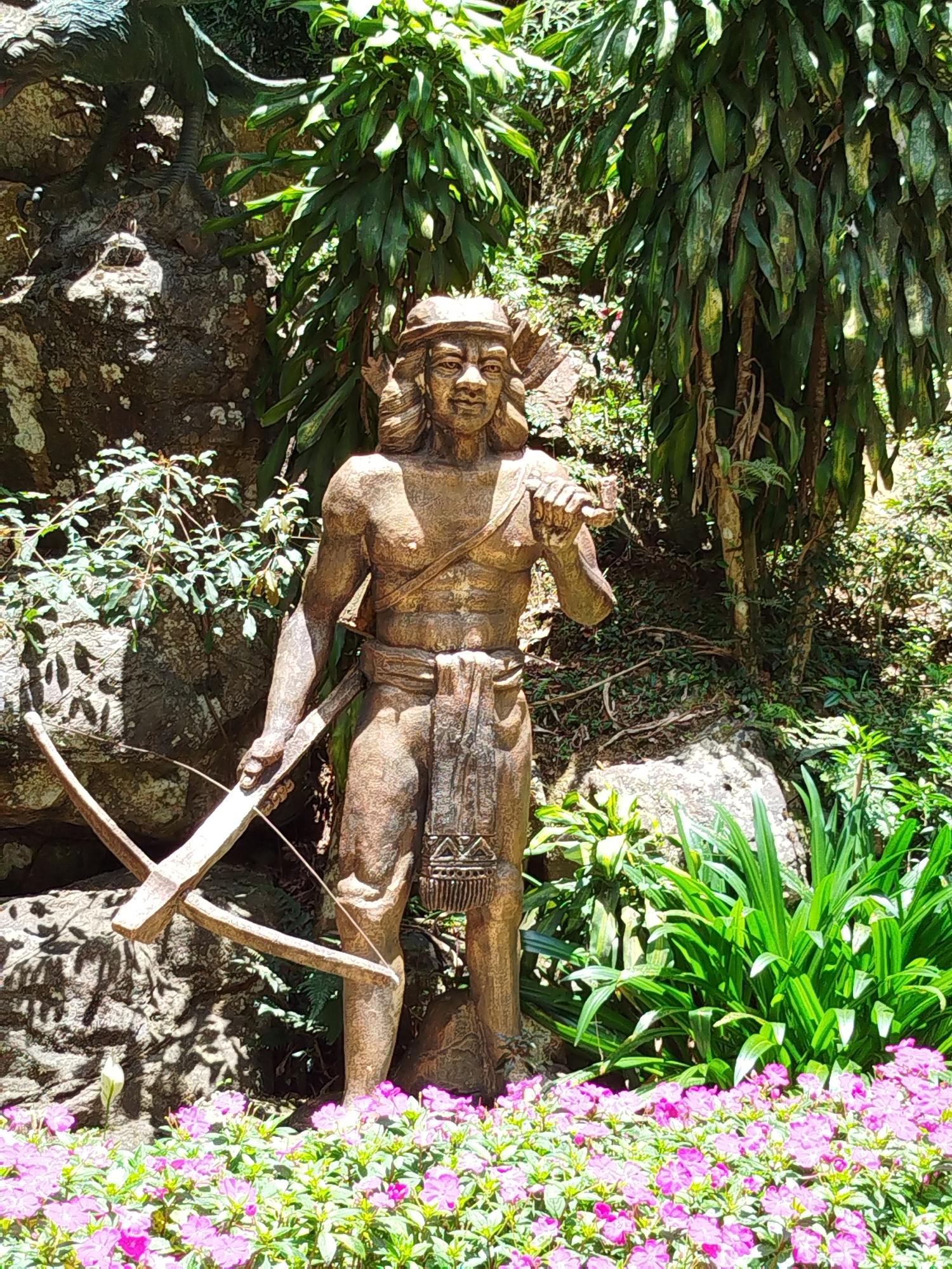 A bronze statue of a male figure stands amid lush vegetation. The statue depicts a man wearing a headdress and a loincloth-like garment, holding a crossbow. The backdrop includes a rock face, various plants, and a bed of pink flowers at the base of the statue. The location is Da Lat, Vietnam.