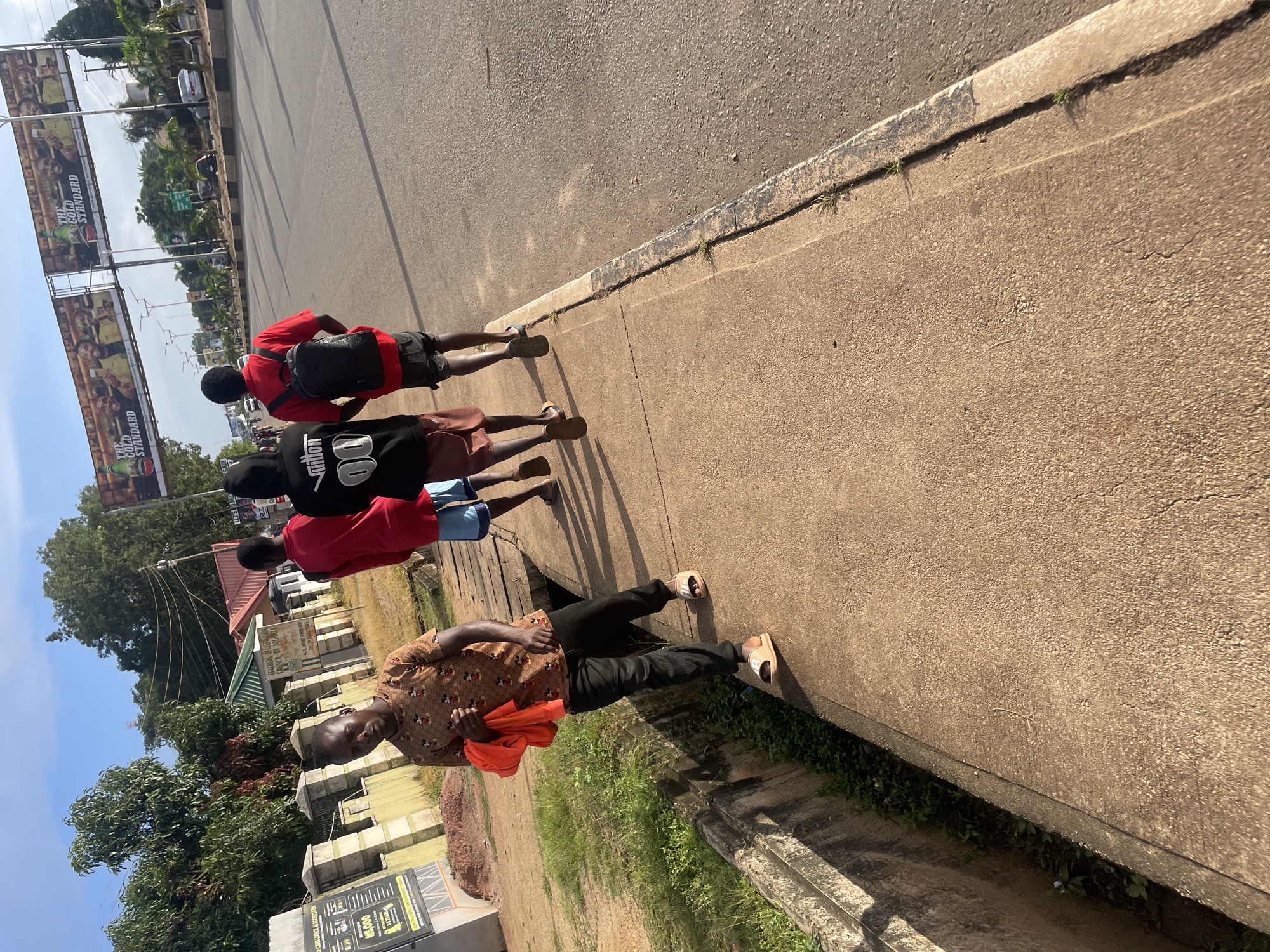 The image captures a scene on a paved sidewalk next to a road in Jos, Nigeria. Four individuals, appearing to be young men, are walking away from the camera along the sidewalk. The person closest to the camera, on the left, is wearing a brown t-shirt with a Mickey Mouse pattern, black pants, and light-colored slide sandals. He is holding a bright orange garment in his right hand. The other three individuals are walking ahead of him. The second person is wearing a red t-shirt and blue shorts. The third person is wearing a black hoodie with "autism 00" printed on the back and brown shorts. He also has a black backpack on. The fourth person, partially visible in the distance, is wearing a red t-shirt and dark shorts.
To the left of the sidewalk, there is a concrete drainage ditch filled with green vegetation. Beyond the ditch, there is a low wall, some trees, and buildings, including one with a sign that appears to read "TEACHER EDUCATION..." and "COUNCIL OF NIGERIA". Further in the background, across the road, is a billboard with advertisements featuring men holding bottles and the text "THE GOLD STANDARD". The road itself is paved, with markings and streetlights visible. The sky is blue with some clouds, suggesting daytime. The overall environment appears to be a relatively urban or semi-urban area.