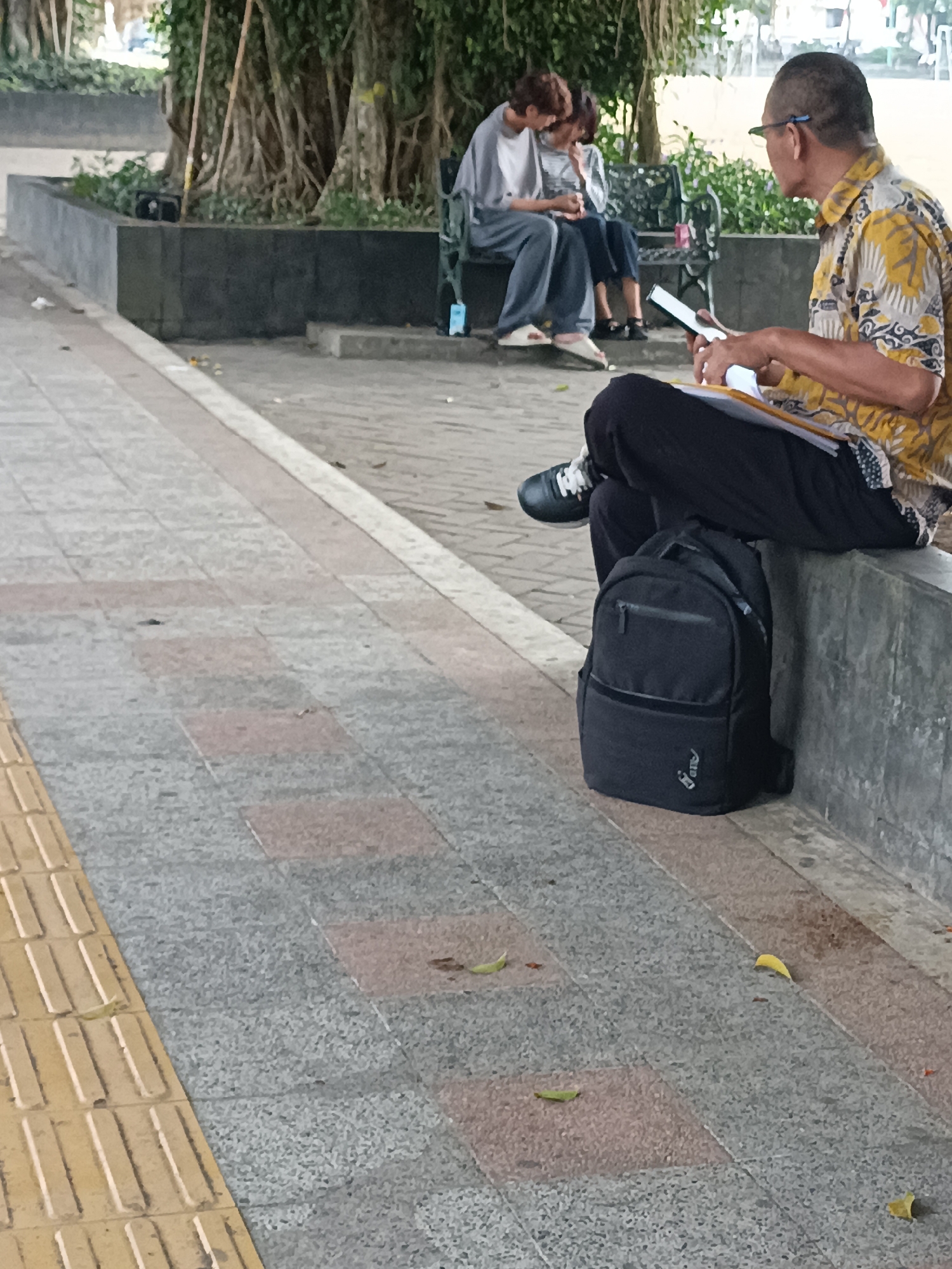 In Sleman, Indonesia, a man in a yellow batik shirt is seated on a concrete ledge, engrossed in a book. Beside him rests a black backpack. Further back, two individuals are seated on a bench, appearing to be engaged in a shared activity, possibly reading or interacting with a device. The scene unfolds on a paved walkway, bordered by a planter box with a large tree. The ground is a mix of grey and pinkish-red paving stones, with a yellow tactile paving strip along one edge. Scattered leaves suggest an outdoor, potentially park-like setting.