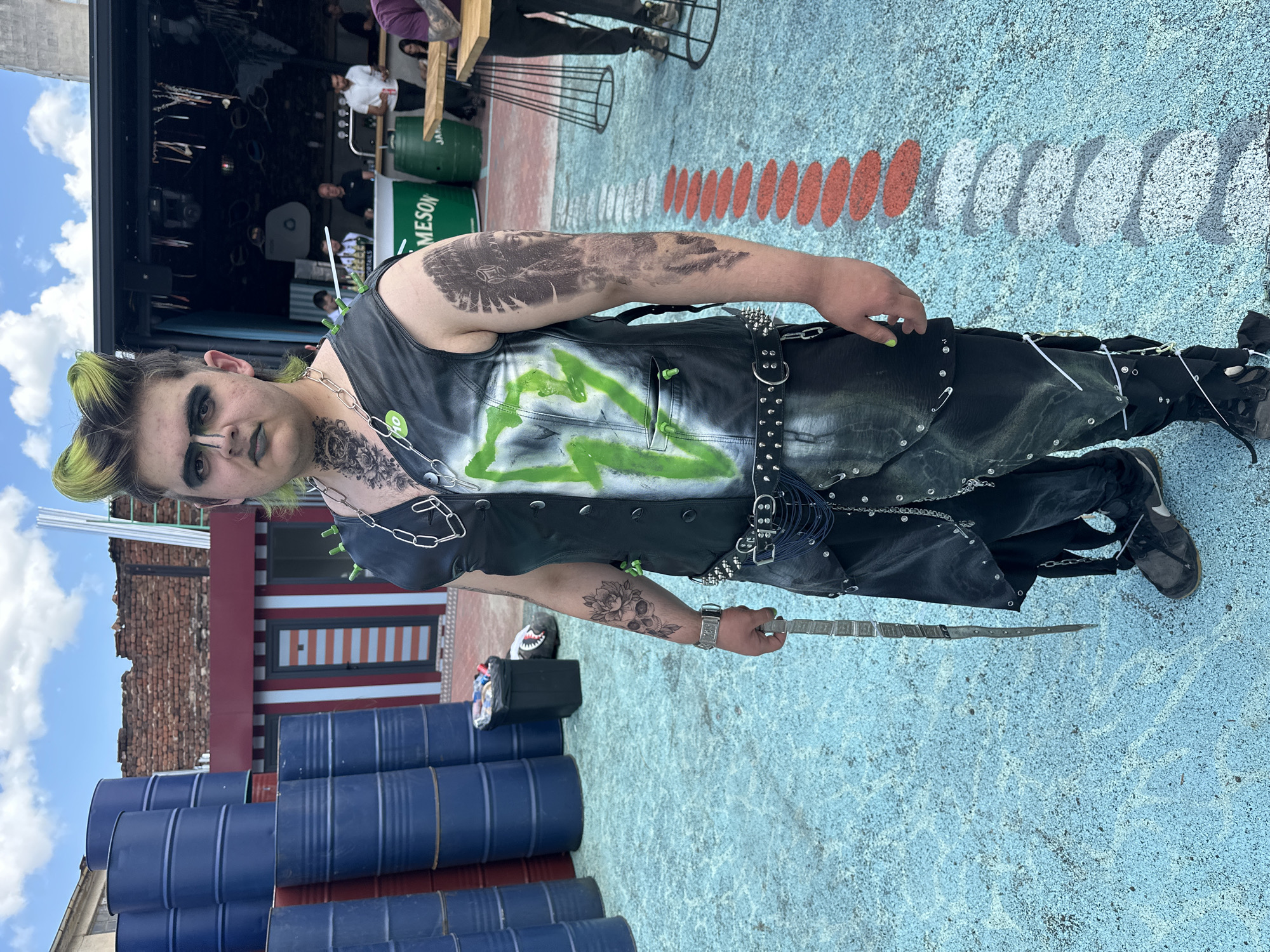 The image depicts a young adult, possibly male, with a distinctive punk aesthetic, standing in an outdoor paved area in Sofia, Bulgaria. The individual occupies the foreground, facing the camera directly. **Person:** The person has bright green hair styled into two prominent upward spikes and dark roots. Their face is made up with heavy black eyeliner creating a winged effect, a black line across the bridge of the nose connecting the eyebrows, and dark greenish-grey lipstick. They wear a black sleeveless vest, possibly leather, with a large, bright green spray-painted lightning bolt design on the left side. A silver chain is draped over the left shoulder, and a small green pin is attached to the upper left chest. Around the waist, a studded black belt with a large buckle secures a layered garment that resembles a skirt over trousers, both black, embellished with silver studs, chains, safety pins, and white zip ties. Dark sneakers with white accents are worn on the feet. The person's left hand, painted with neon green fingernails, holds a long, slender, metallic object resembling a ceremonial dagger or sword, held upright. A silver-colored watch is visible on the left wrist. Visible tattoos include an intricate black floral and skull design on the left forearm, a large black tattoo depicting an Indigenous American chief's head and headdress on the right upper arm, and black floral/tribal patterns on the neck and collarbone. **Environment:** The ground is a light blue/teal, speckled, textured surface, possibly painted concrete or asphalt. A decorative pathway consisting of alternating oval shapes, painted orange-red and white/grey, extends from the right foreground towards the background. On the left side of the background, several stacks of blue industrial barrels are visible, some appearing worn or rusted, with hints of red barrels underneath. Behind these barrels, a partial brick wall of an older building is present, and a smaller structure with a red facade and a vertically striped door (red and white/grey) stands next to it. A black trash bin is located near the barrels. On the right side of the background, an outdoor bar area is set up under a dark canopy. A green counter displaying the "JAMESON" logo is visible, flanked by two green barrels. Several individuals are present in this area, some appearing to be staff and others patrons, standing or sitting at high-top wooden tables with black metal bases. The sky above is blue