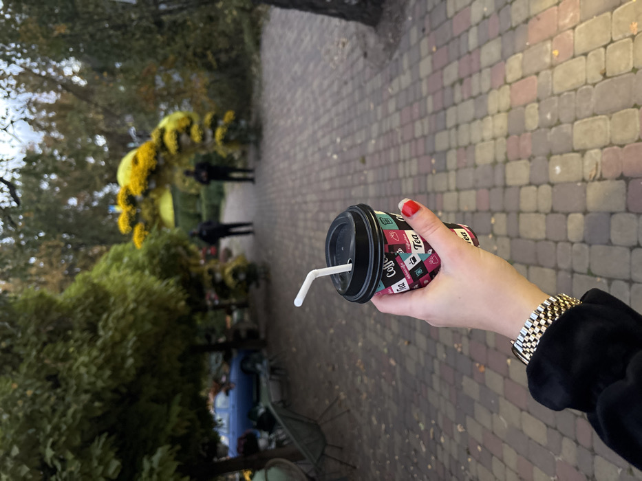 A close-up, first-person perspective image depicts a fair-skinned human hand holding a multi-colored disposable cup with a black lid and white bent straw, positioned over a paved pathway in Lisne, Ukraine. The hand, adorned with red nail polish on visible fingers and a gold-toned metal link bracelet on the wrist, wears a dark, possibly black, sleeve. The cup features "Coffee" and "Tea" text along with various beverage-related icons.
The background, out of focus, shows an outdoor scene with a path constructed from interlocking multi-toned pavers (reddish, brown, gray). To the left, a blue car is partially visible behind lush green trees, alongside outdoor seating arrangements consisting of tables and chairs. Further down the path, a prominent decorative archway made of yellow flowers, likely chrysanthemums, spans the walkway. Beyond this archway, two indistinct, dark-clad figures stand on the path. Faintly visible in the distant foliage behind the archway are elements resembling a blue and yellow flag. The environment suggests a park, garden, or pedestrian area with mature trees and designed floral elements.