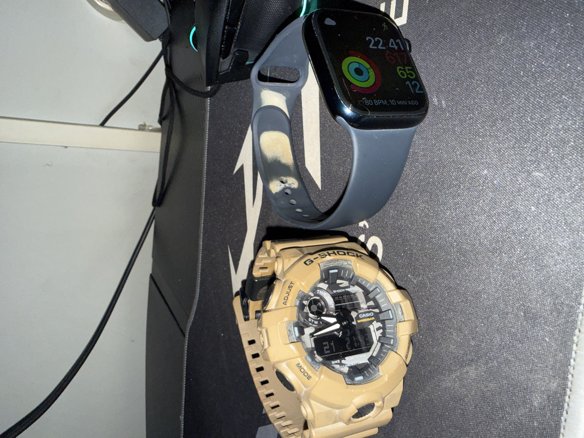 The image presents a close-up view of two wristwatches and a black object, possibly headphones, arranged on a dark, textured surface. The upper watch is an Apple Watch with a dark blue strap, displaying the time as 22:41 and various health metrics. The strap has a noticeable worn or discolored patch. Below it is a G-Shock watch in a light brown or khaki color, showing the time as 21:28 on its digital display. A black cable extends from the side of the image, potentially connected to the black object at the top left. The environment appears to be a desk or tabletop. There is no evidence to place this scene in Shumen, Bulgaria.