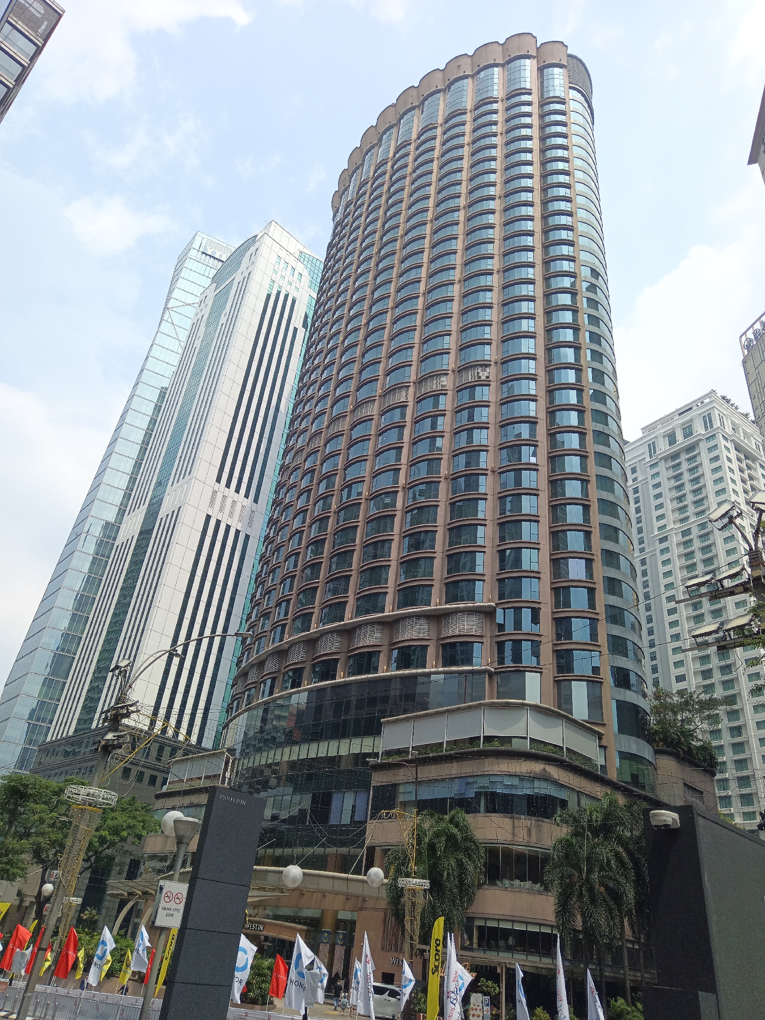 The image shows a low-angle shot of several modern skyscrapers in Kuala Lumpur, Malaysia, under a bright, overcast sky. The most prominent building, a tall, curved tower with a dark brown facade and numerous reflective windows, dominates the center of the frame. To its left, a sleek, glass-fronted skyscraper with a diagonal design stands tall, featuring vertical white accents. In the background, another lighter-colored, rectangular skyscraper with many windows is visible, and further to the right, a section of a building with a more traditional, grid-like window pattern can be seen. At the base of the main curved building, there are visible signs, including one that reads "Pavilion" and another with the "WESTIN" logo, indicating the location is likely the Pavilion Kuala Lumpur shopping mall and its connected hotel. Several flags, predominantly white and red, are displayed on poles at street level in front of the buildings. Palm trees and other greenery are also present, suggesting an urban environment with landscaping. There are no visible people in the frame. The overall impression is one of a bustling, contemporary urban landscape.