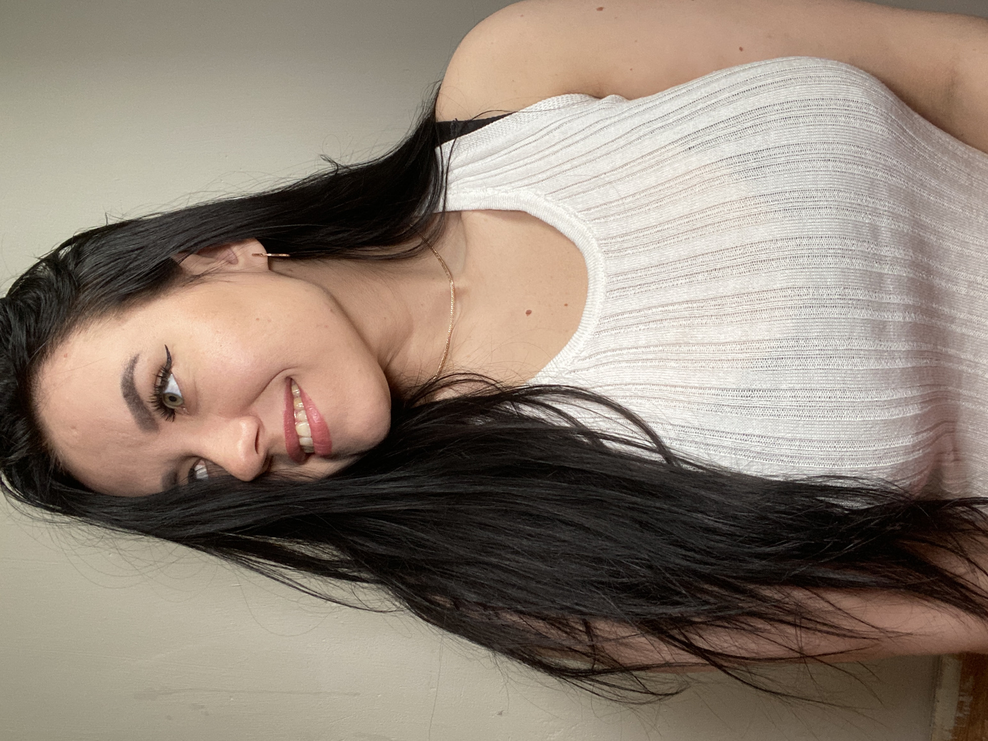 The image is a rotated portrait capturing a young woman with a warm, smiling expression. She has long, dark, straight hair cascading over her shoulders and chest. Her face features prominent dark eyebrows, winged eyeliner accentuating her light green or hazel eyes, and a broad smile revealing her upper teeth. Her lips are painted with a light pink or nude lipstick.
She is wearing a light-colored, possibly white or off-white, ribbed sleeveless top. A black bra strap is visible on her left shoulder. Accessories include a delicate gold chain necklace around her neck and a small gold hoop earring in her visible ear. The background is a plain, light-colored wall, suggesting an indoor setting with simple, diffuse lighting. There is no visible text. The activity is a posed portrait, conveying an emotion of happiness and friendliness. Based on the provided context, the scene is located in Chaikovsky, Russia.