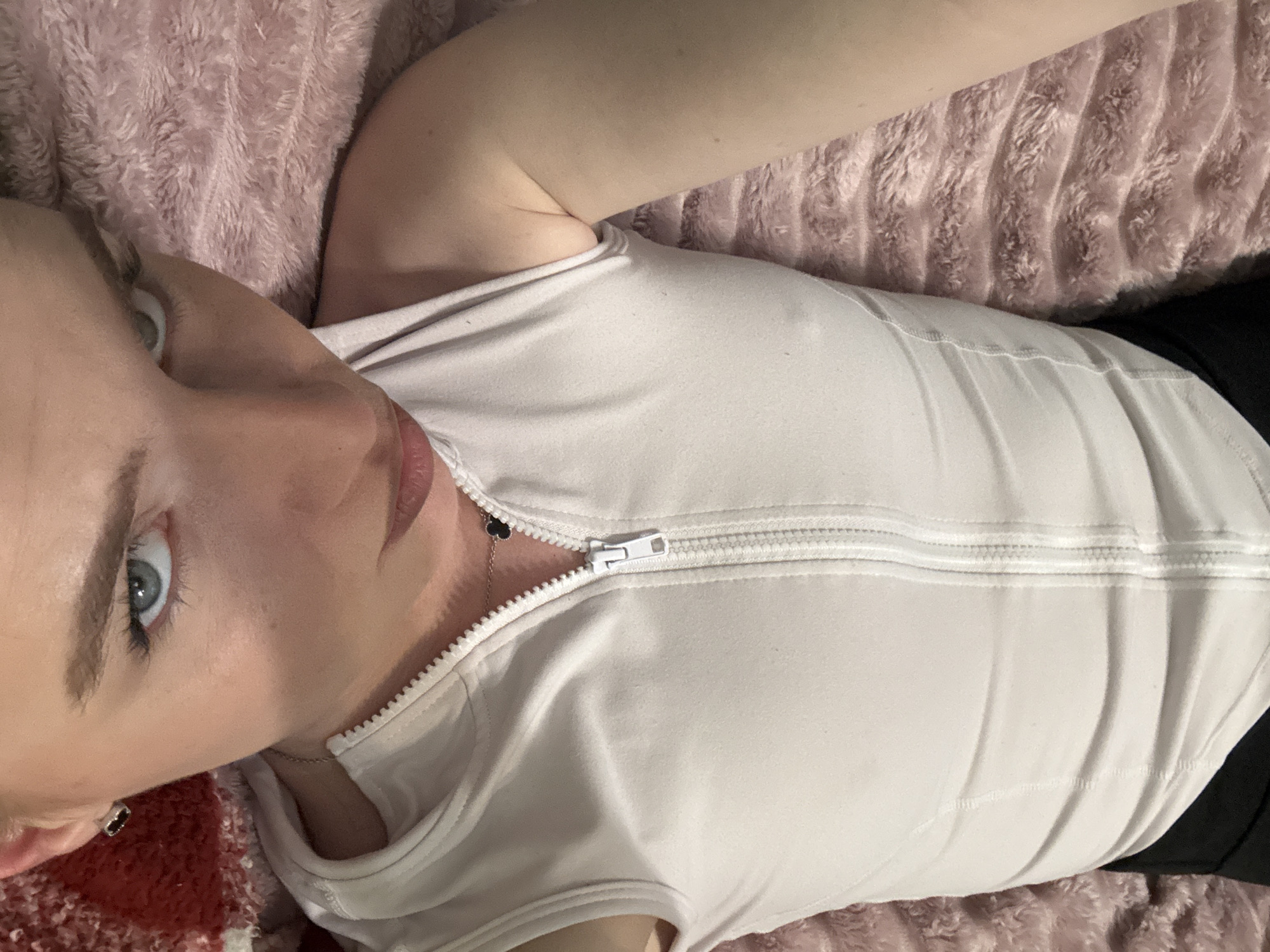 A woman lies on a pink and red textured surface, possibly a bed. She is wearing a white, zippered top and black bottoms. A delicate necklace with a black charm is visible around her neck. The location is not discernible from the image.