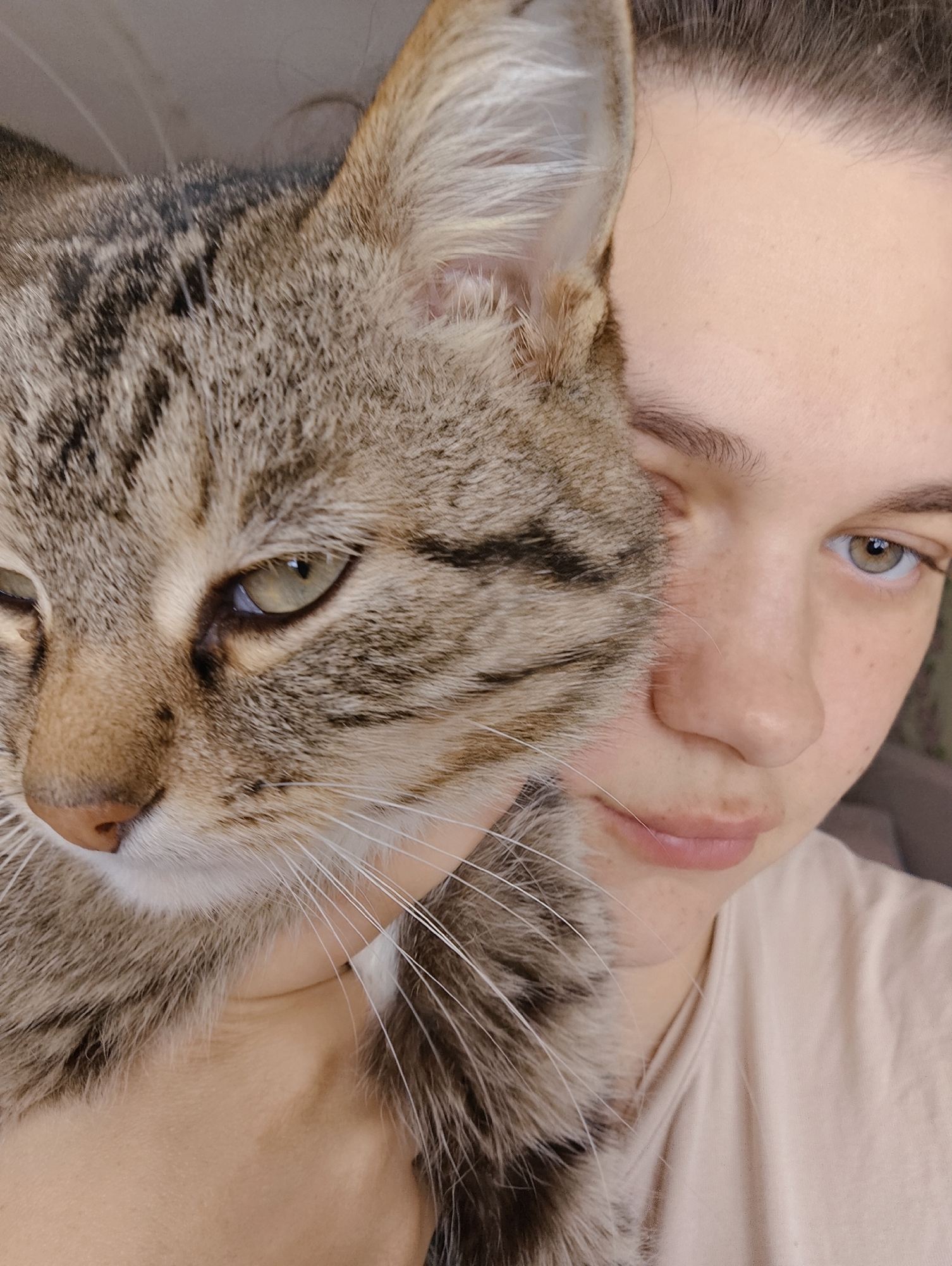 A fair-skinned young adult female with brown hair pulled back is holding a brown tabby cat closely against her face. The cat's face fills the left side of the frame, while the woman's face occupies the right side. The woman is looking slightly downward, her expression soft. The cat is also looking downward, appearing somewhat melancholic. The background is out of focus. There is no indication of the city Уральское, Russia.