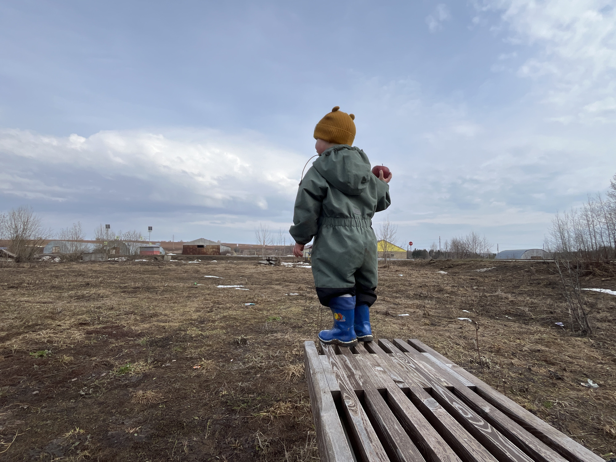 A child stands on a wooden bench in an outdoor location in Павловка, Russia. The child wears a yellow knit hat, a green jumpsuit, and blue boots. The child is holding a red apple. The ground is covered in brown grass and dirt. There are buildings in the background and a cloudy sky overhead.