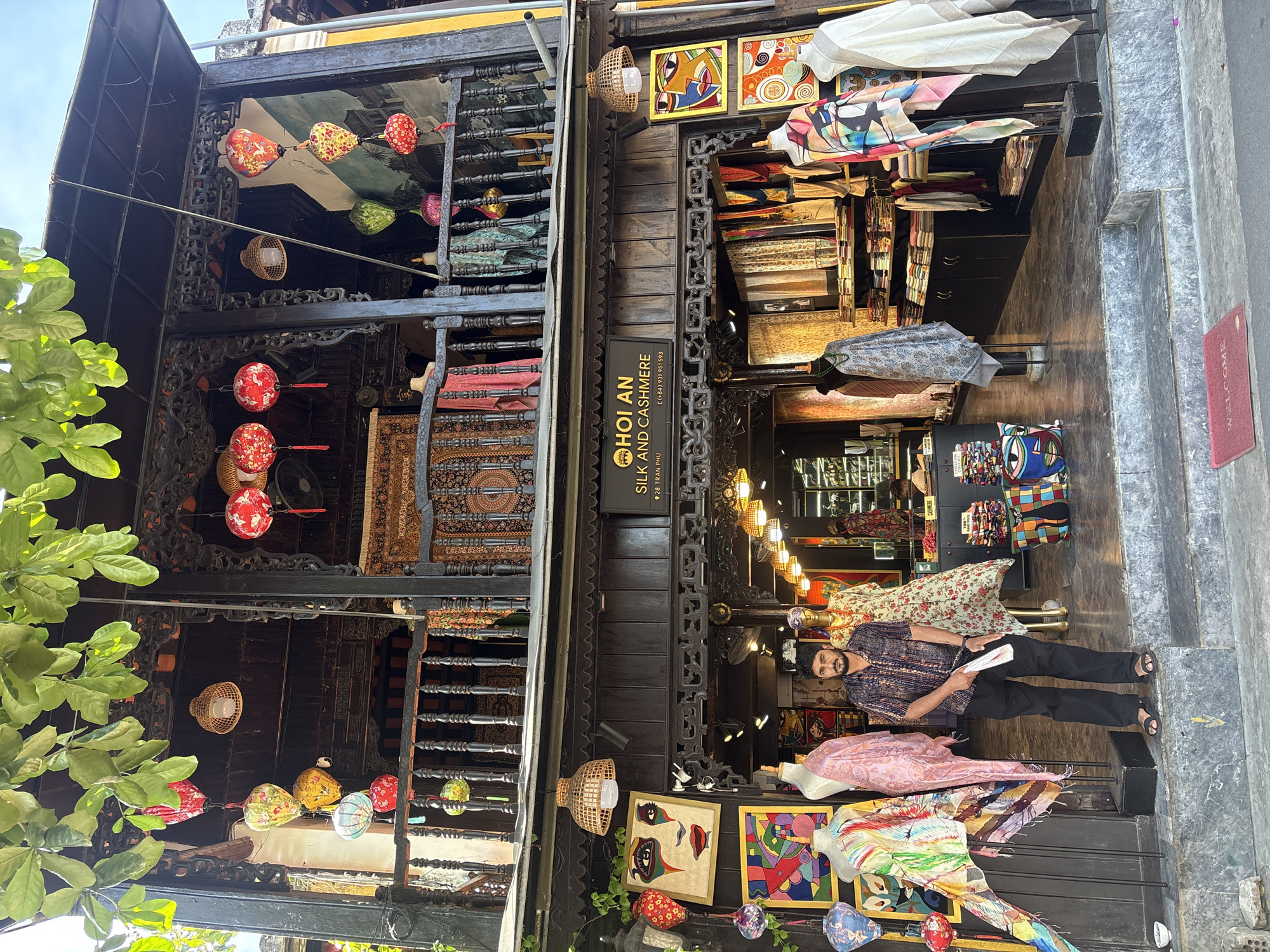 The image shows the exterior of a shop in Hoi An, Vietnam, that sells silk and cashmere. The shop is a two-story building with dark wood accents and a balcony decorated with colorful lanterns. In front of the shop, there are several mannequins displaying scarves and other textile items. A man stands on the steps leading into the shop, holding a white piece of paper. The shop's name, "HOI AN SILK AND CASHMERE," is visible on a sign above the entrance. Artwork, including paintings of faces and abstract designs, is displayed on the exterior walls. Lush green foliage hangs from above, partially obscuring the upper part of the building. The ground in front of the shop is paved with stone, and a red "Welcome" mat is placed on the ground. The overall impression is of a vibrant and traditional shop in a bustling tourist area.
