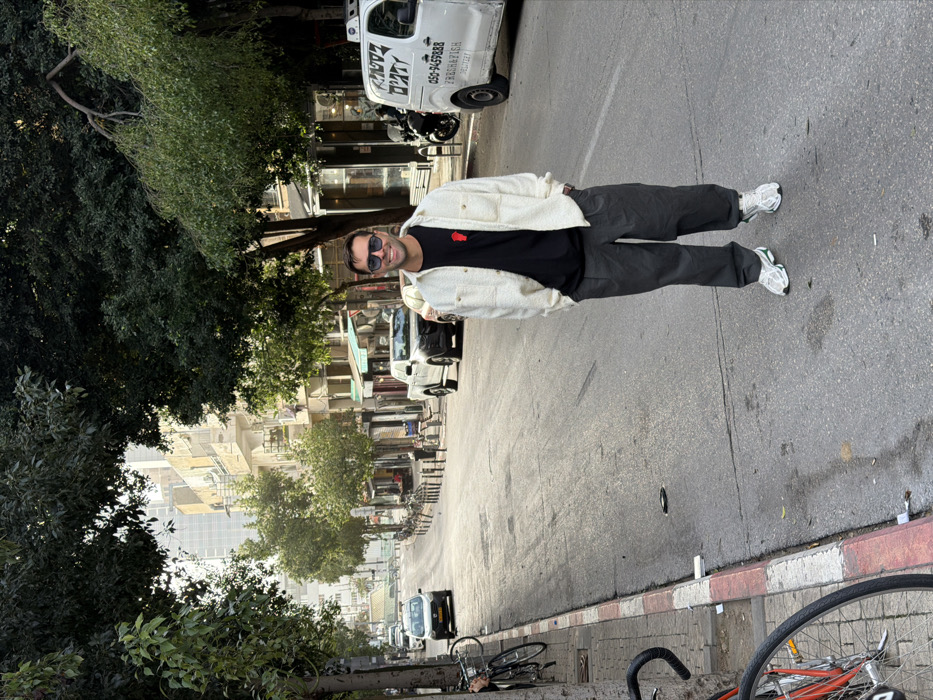 The image displays a male individual standing on an asphalt street in Tel-Aviv, Israel, under natural daylight. He is positioned centrally, facing forward, with a slight smile, wearing dark sunglasses. His attire consists of a cream-colored textured overshirt worn open over a black t-shirt with a small red motif on the left chest, dark grey pleated trousers, and white athletic shoes with green accents. His hands are in the pockets of his overshirt.
The street features a red and white striped curb along the left edge, where a sidewalk is visible with several bicycles chained to trees. Multiple mature trees line both sides of the street, creating an overhead canopy. In the background, various multi-story buildings line the street, displaying different architectural styles. Vehicles are present: a white compact car is visible on the left side of the street, followed by other vehicles receding into the distance. On the right side, a black pickup truck is parked, followed by a white delivery van. The delivery van has Hebrew text and "FRESH FISH DELIVERY" written on its side, along with a phone number (050-9459XXX), identifying it as "Radotz Fresh Fish Delivery," which operates in Tel-Aviv. Bollards are visible along the right side of the street. The street itself appears to gently incline upwards in the background.