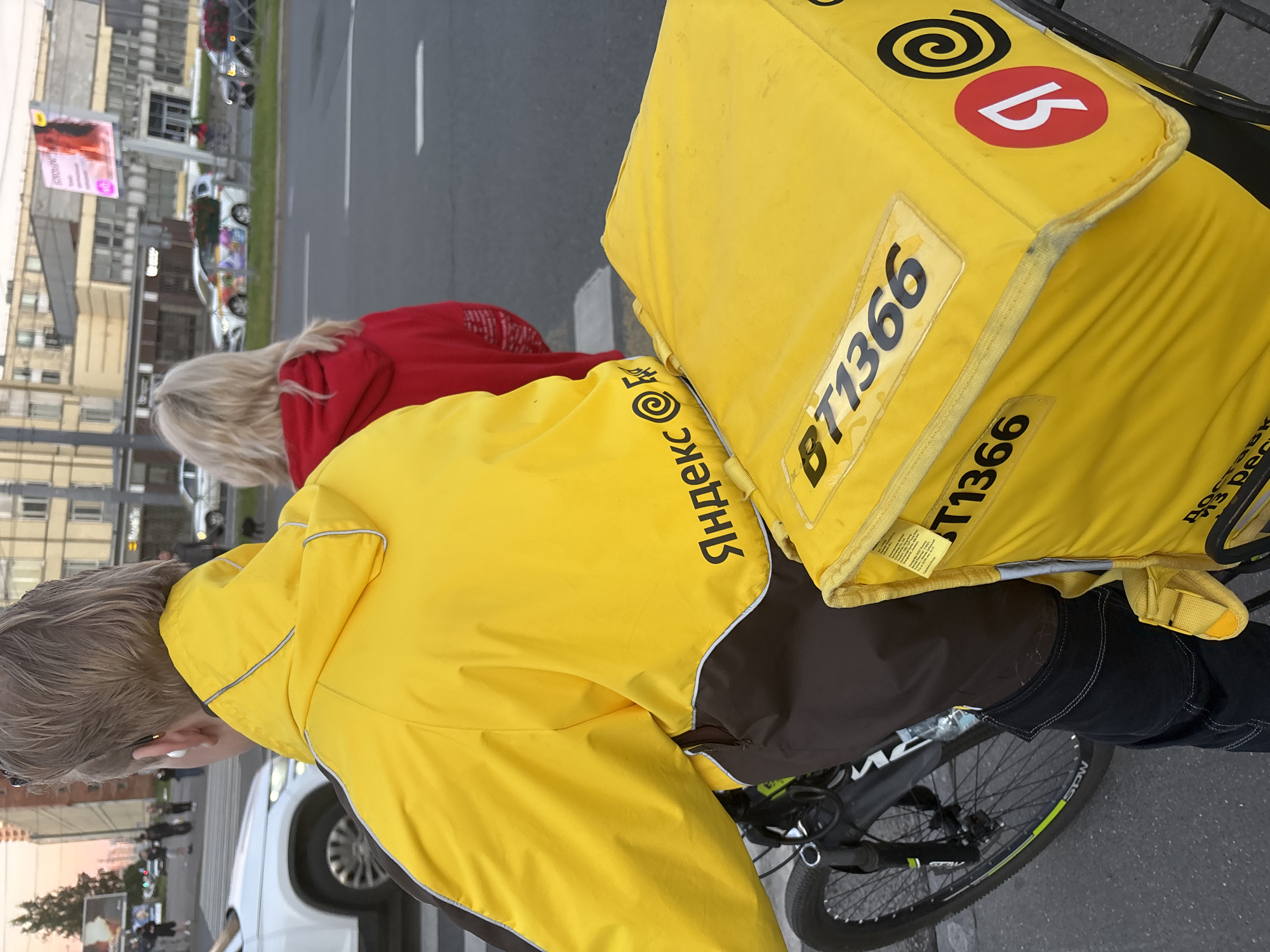 In Sinyavino, Russia, a person wearing a yellow jacket with brown accents is riding a bicycle. The jacket has a logo that appears to be "Яндекс." Attached to the back of the bicycle is a large, yellow insulated bag, also featuring the "Яндекс" logo and a number "BT1366". To the right of the cyclist, another person wearing a red hooded sweatshirt is visible, facing away from the camera. The scene takes place on a street with a road and sidewalk, with buildings and other vehicles visible in the background. The lighting suggests it may be late afternoon or early evening.