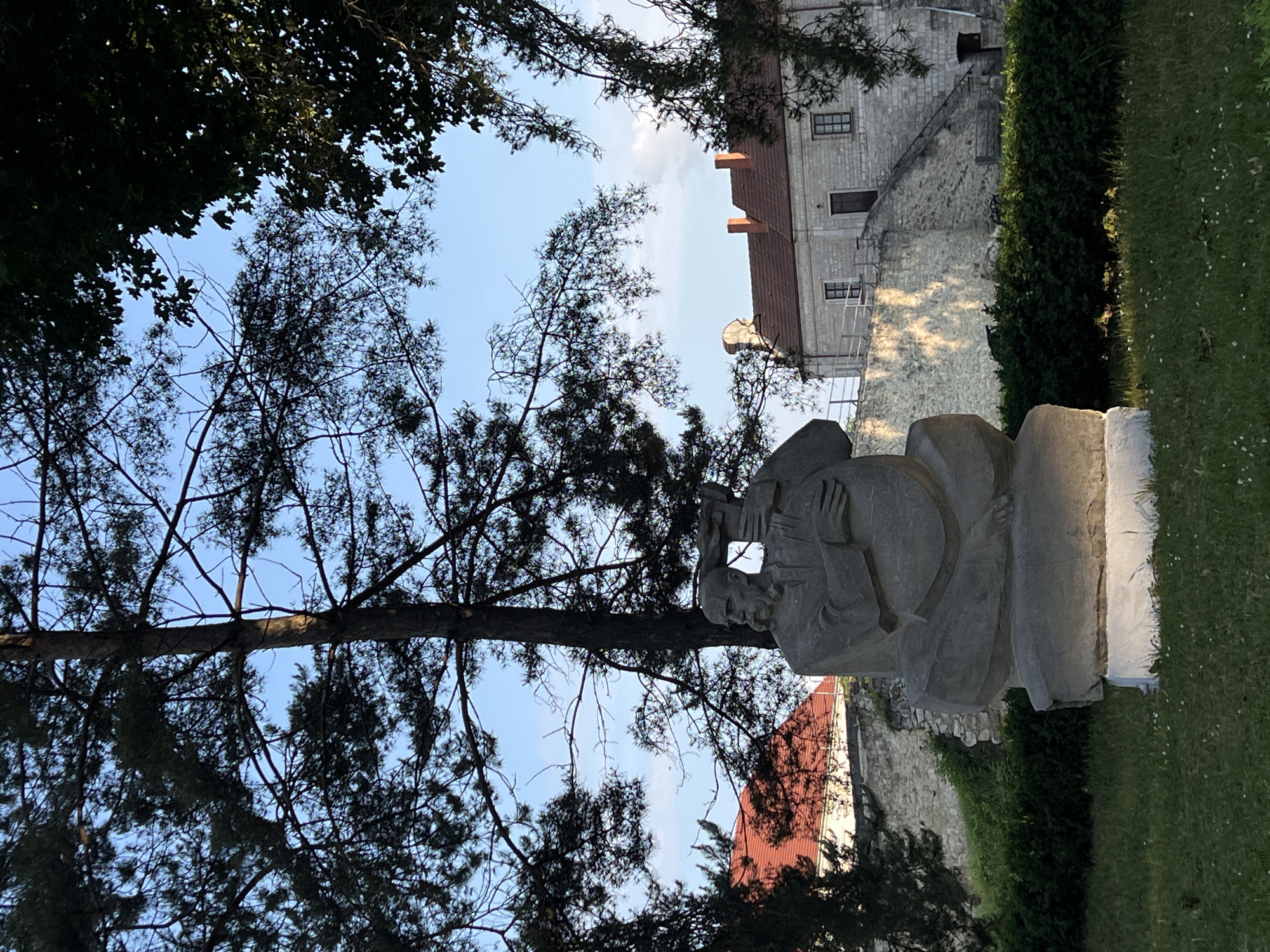 The image, viewed upright, features a light-colored stone statue of a male figure, possibly an older man with a beard, seated or in a reclining pose, holding an object resembling a book or tablet. The statue is positioned in a green, grassy area, likely a park or garden.
The scene is framed by numerous tree branches and foliage, silhouetted against a bright, clear blue sky, suggesting ample natural light. In the background, a historic stone building with a reddish-brown tiled roof is visible. This building, constructed from large stone blocks, features several dark-framed windows and appears to be part of a larger complex or fortification. Adjacent to it, a tall, neatly trimmed green hedge runs alongside a stone wall, further suggesting a landscaped outdoor setting.
The lighting indicates a sunny day, likely during the late afternoon or early morning, with soft, directional light illuminating parts of the building and the ground. No discernible activity is taking place, and there is no visible text on the statue or any structures. The architectural style aligns with historical sites, consistent with the provided location context of Zbarazh, Ukraine.