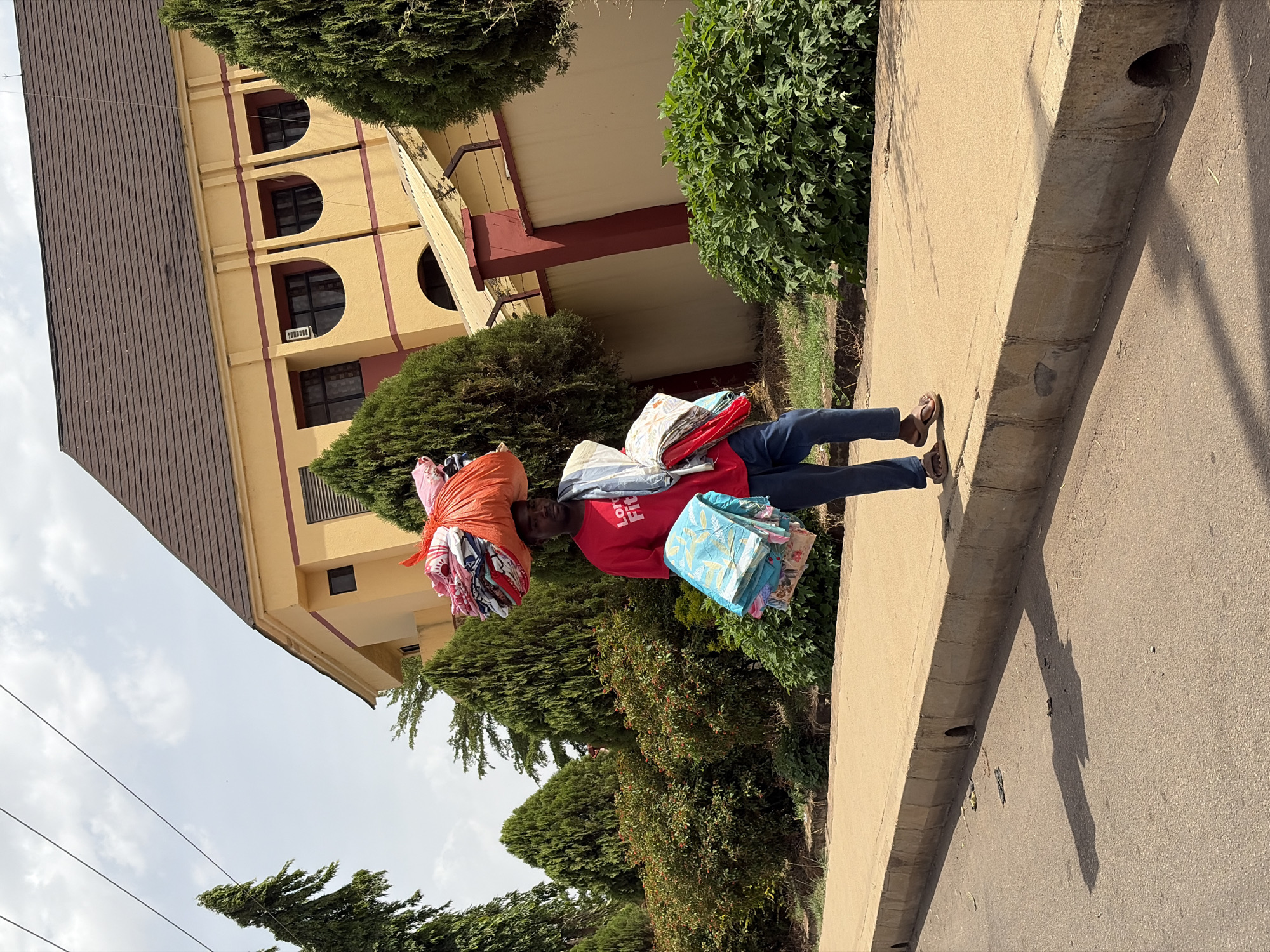The original image is rotated 90 degrees clockwise. Describing it in an upright orientation, the scene captures an individual walking along a sidewalk next to a street in Rafin Par, Nigeria, during the daytime.
The subject is a man wearing a red t-shirt, blue trousers, and light-colored sandals. He is actively engaged in carrying a significant amount of textile goods. A large, orange-wrapped bundle of patterned fabrics is balanced on his head, while smaller bundles of brightly colored and patterned clothes are carried in his arms: one in his left arm and another, primarily blue, in his right arm. On his red t-shirt, the text "Long Life Fit" is visible across the chest.
To the man's left is a multi-story building featuring a yellow facade with distinct maroon banding and a dark brown, sloped roof. The building has several windows with arched tops, and at least one rectangular window with an external air conditioning unit. Dense green bushes and trees line the sidewalk, separating it from the building. The sidewalk itself is concrete, and to the man's right, there is a paved asphalt road. Long shadows are cast on the road, suggesting a bright sun even under a partly cloudy sky. Overhead, utility lines are visible against the sky. The image portrays a slice of daily life, likely a street vendor or hawker, typical of the region.