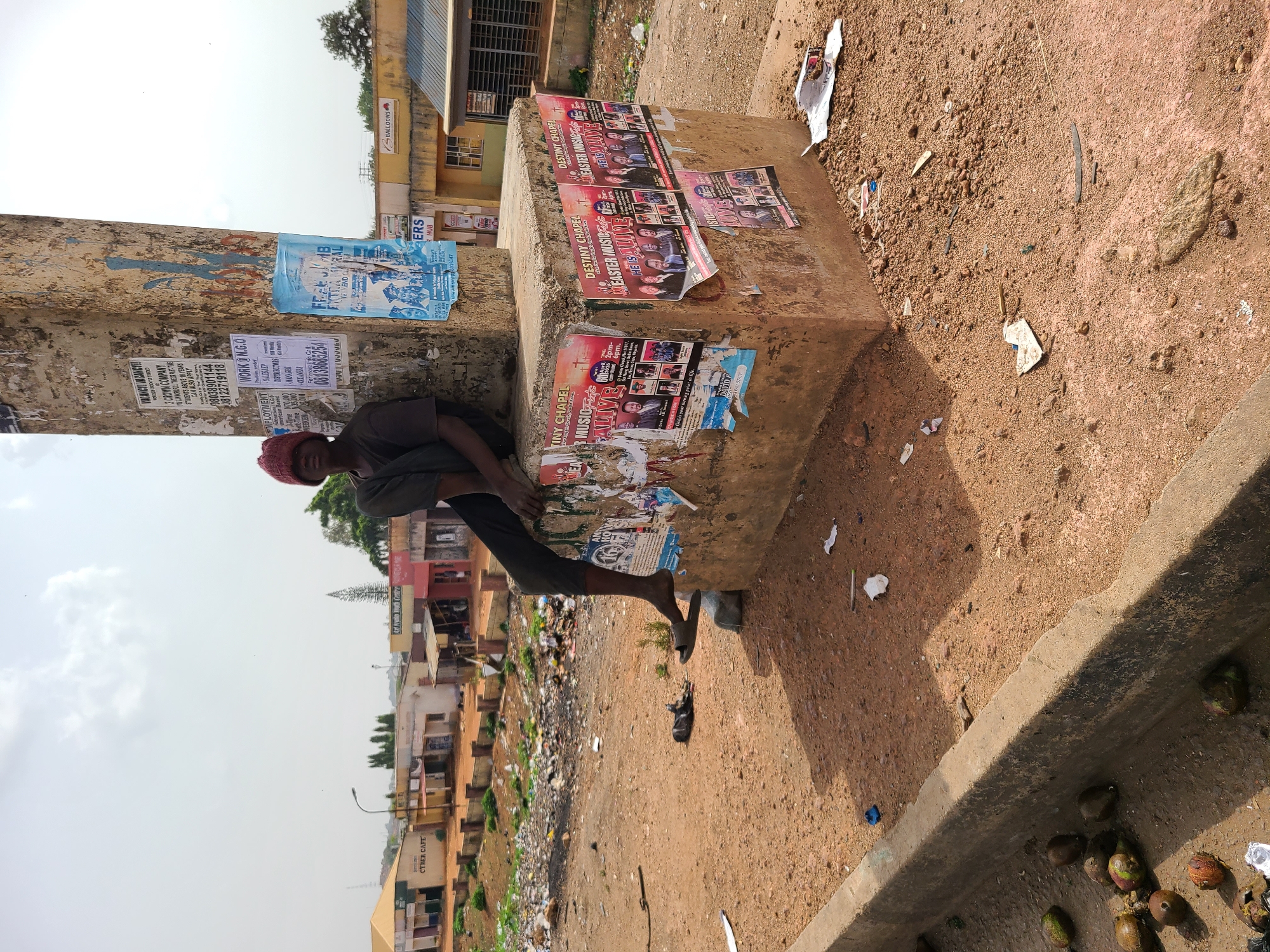 An outdoor, daytime shot, likely taken in Jos, Nigeria, shows a man leaning against a concrete pillar in what appears to be an urban or semi-urban setting. The image is oriented vertically (originally rotated 90 degrees counter-clockwise).
The main subject is a man, wearing a red cap and dark clothing, who is resting against the left side of a weathered concrete pillar. His posture suggests he is either taking a break or waiting. His footwear consists of simple slip-on sandals.
The environment is characterized by a reddish-brown dirt ground, scattered with debris including small stones, pieces of paper, plastic, and discarded fruit shells (possibly coconuts or similar). In the background, beyond the man and the pillar, there is a visible street or path leading to multiple simple, multi-story buildings. Some buildings feature corrugated metal roofs. The sky overhead is bright and partly cloudy, indicating daytime, with strong shadows suggesting direct sunlight.
The concrete pillar and a connecting concrete ledge or bench are heavily adorned with various posters and flyers, many of which are torn or layered. Visible text includes advertisements for "VACANCY! VACANCY!" at "3 CROWN COMPANY" for "STUDENTS ABOVE 18 YEARS," providing phone numbers "08038901144" and "08122719118." Other employment ads mention "WORK@N.G.O" for "SALES REP" and "MANAGER" positions, with potential earnings of "N70,000" for full-time work and another contact number "081360654." Religious flyers for "DESTINY CHAPEL EASTER MUSIC F HE IS ALIVE" and "TINY CHAPEL MUSIC F ALIVE" are also prominent, some noting "2pm-6pm" events. One poster reads, "God is your turning point in life." In the background, a building has a sign that reads "CYBER CAFE." Other faint texts visible are "BALLOONS" and "NUB."
The scene depicts a bustling, lived-in environment, characteristic of a street or communal area in Jos, Nigeria, where public spaces serve as informal bulletin boards.