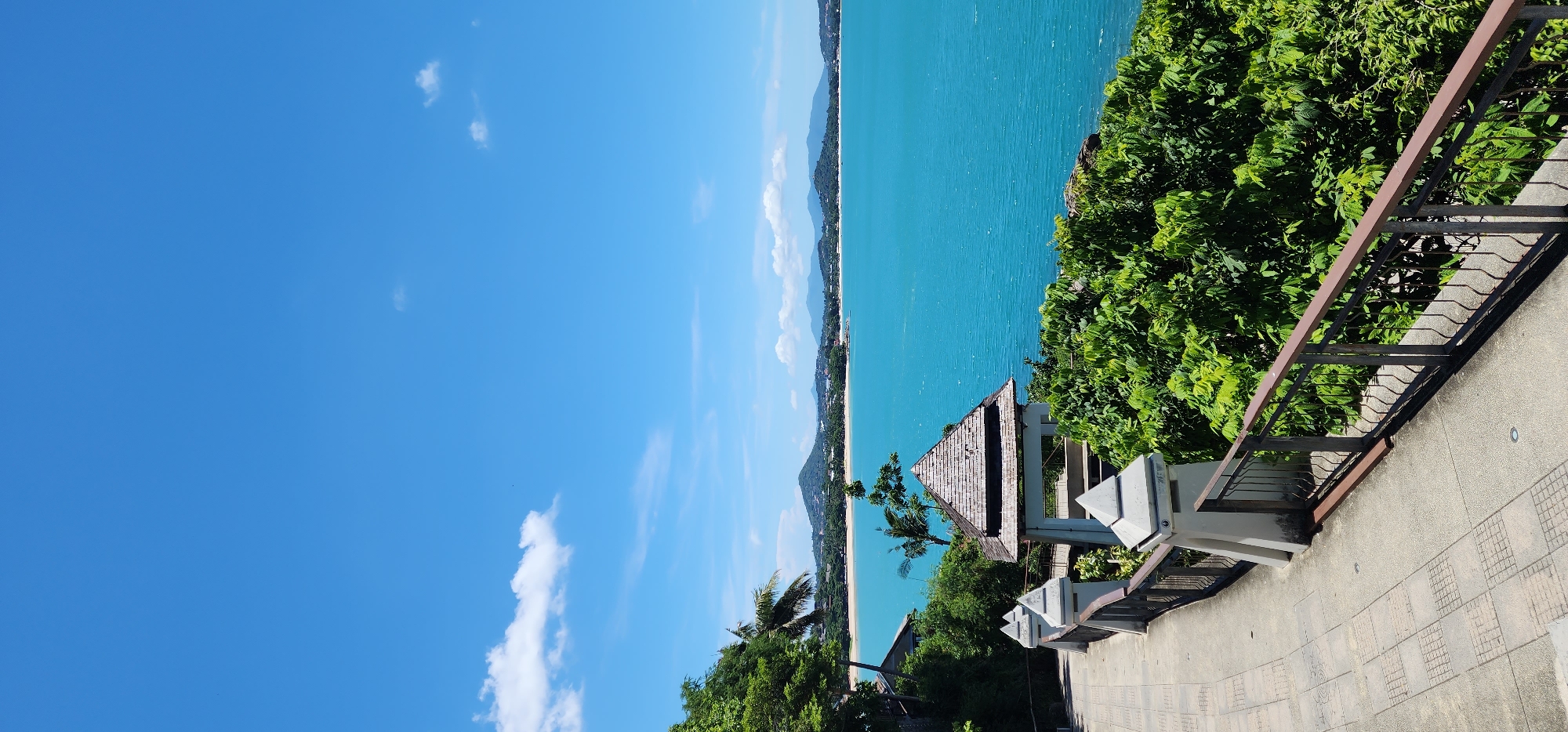 The image depicts a scenic coastal view in Baan Chaweng Noi, Thailand. A paved walkway with decorative railings curves to the left, bordered by lush green vegetation on the right. In the foreground, a small, traditional Thai-style gazebo with a wooden roof stands amidst the foliage. Beyond the greenery, the bright turquoise waters of the sea stretch out to the horizon, meeting a clear blue sky dotted with a few wispy white clouds. In the distance, a long, sandy beach is visible, backed by rolling hills and mountains, suggesting a tropical island landscape. There are no people or animals visible in the image. The scene is bathed in bright daylight, indicating a sunny day.