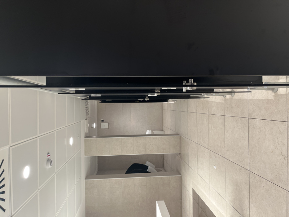 This image depicts a modern, empty public restroom in Dorno, Italy. The perspective is slightly angled, looking towards a row of restroom stalls. The scene is characterized by clean lines and neutral tones.
The main subjects are the restroom stalls, which feature dark, possibly black, partitions with chrome-like hardware for hinges and latches. At least three stalls are visible, with two toilets identifiable in the background. One toilet is within an open stall, and another is partially visible behind a low partition. In the foreground of one stall, a black object, possibly a bag or piece of clothing, lies on the floor.
The left side of the room features white paneled walls with integrated circular lights, illuminating the area. A red fire alarm or smoke detector is mounted on one of these panels, and a ventilation grille is visible further to the left. The floor throughout the restroom is covered in light-colored, square tiles, which also extend partway up the walls in the stall area. On a tiled wall in the background, a white dispenser (possibly for soap or paper towels) is mounted above a small white waste bin. The ceiling appears to be dark, contrasting with the lighter walls and floor.
No people are present, and no specific activity is taking place, suggesting the restroom is currently unoccupied. The lighting is artificial, and there is no indication of the time of day or outdoor weather. No visible text is discernible in the image. The overall impression is one of a clean, well-maintained, and functional public facility.