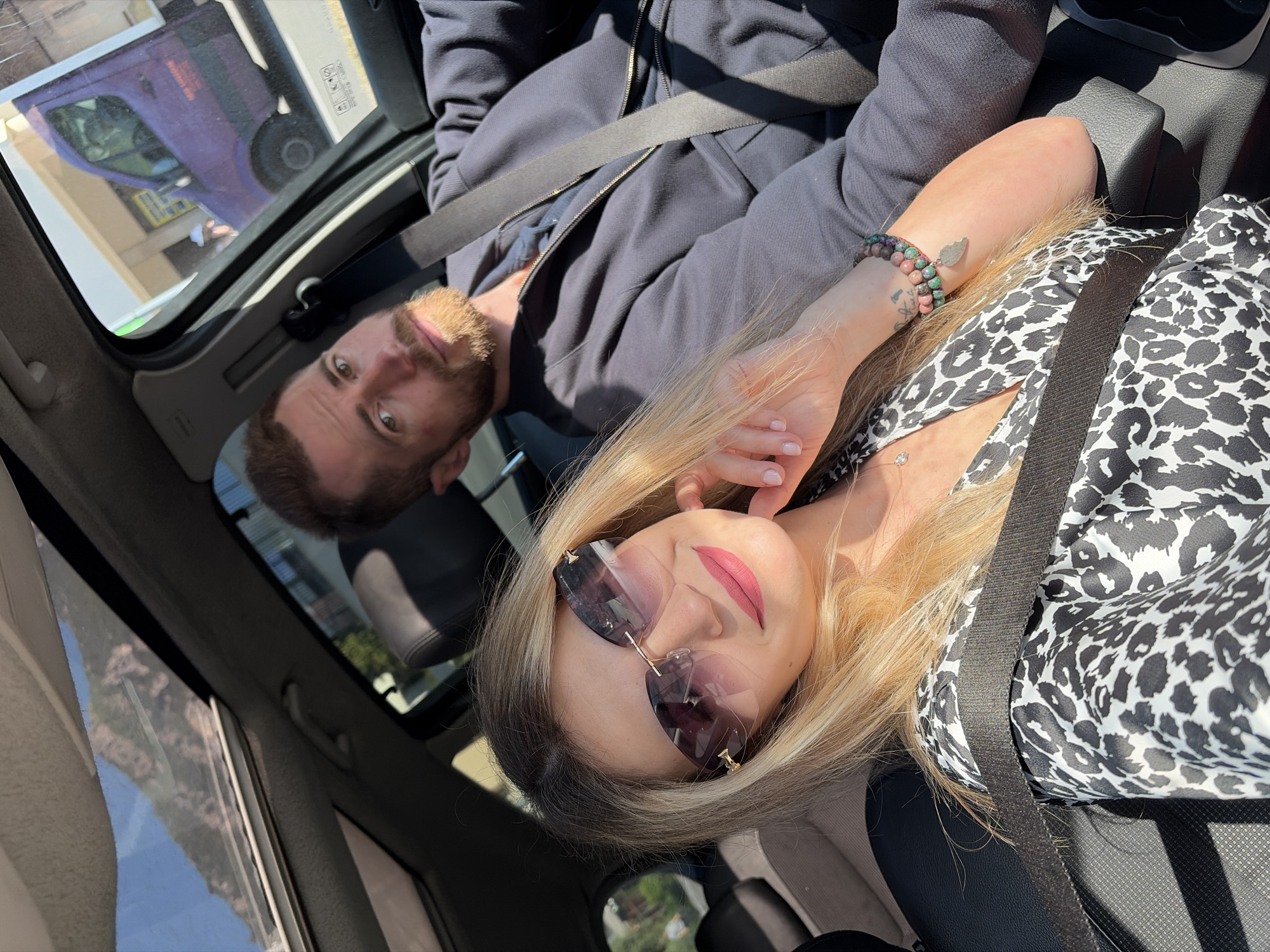 Two people are in a car. A man with a beard is lying back with his seat belt on. A woman with blonde hair and sunglasses is lying next to him, also with her seat belt on. She is wearing a black and white animal print top and has a bracelet on her wrist. The car appears to be driving through the city of Boreti, Montenegro, as indicated by a distant mountain view visible through the car's window. A purple van is visible in the sunroof's reflection.