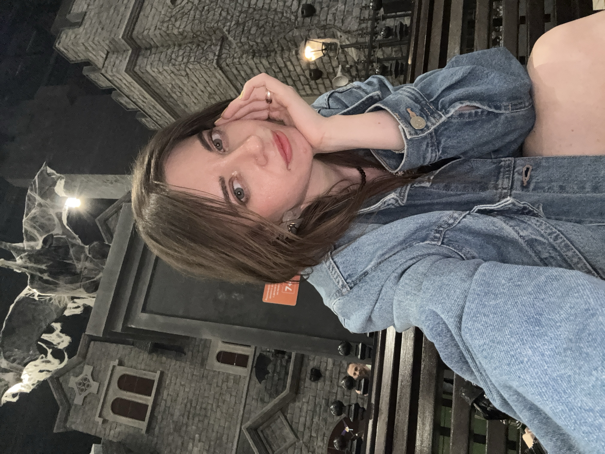 A young woman with brown hair is posed in a selfie. She has fair skin and is wearing a denim jacket. Her face is tilted slightly toward the camera with her hand near her cheek. She is wearing jewelry and has red lipstick on. The background appears to be part of a dark ride attraction or themed environment. The backdrop includes details suggesting a gothic or spooky setting, with what appears to be model buildings and what resembles spiderwebs and a lamppost with a yellow light. There is a wooden bench in the foreground. Given the details in the background, this location appears to be inside the Dream Island theme park in Moscow, Russia.
