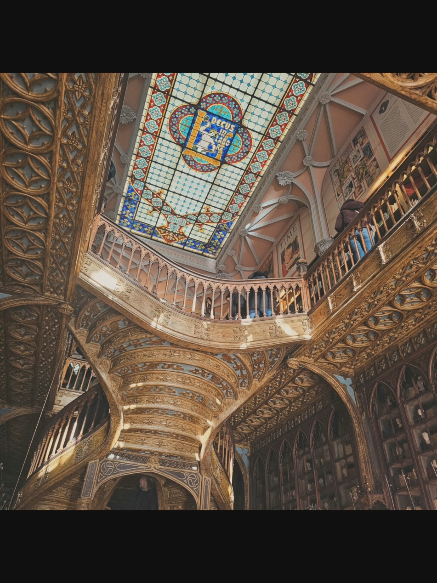 An ornate library interior with a stained glass ceiling and intricate wooden detailing. The ceiling features a central stained glass panel with a blue and gold geometric design and a depiction of a figure. A grand, curved wooden staircase with a balustrade dominates the foreground, leading up to a mezzanine level. The mezzanine also has a wooden balustrade and is lined with bookshelves. Several people are visible on the mezzanine, some browsing shelves and others walking along the balcony. The walls are adorned with framed pictures and decorative architectural elements. The overall impression is one of grandeur and historical significance, typical of a well-preserved old library.