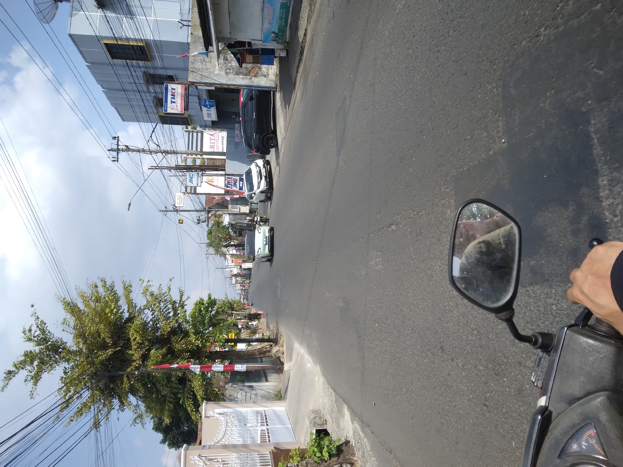 The image captures a forward-facing view from a motorcycle in Mersi, Indonesia, on a paved street lined with buildings and trees under a partly cloudy sky. On the left, a white gate and a beige fence are visible, along with a tall tree adorned with red and white Indonesian flags. The street itself is asphalt, with a visible white line marking the lane. Several vehicles are present: a white car in the middle distance, a black SUV parked on the right, and another white car further down the road. Buildings line the right side of the street, displaying various signs, including "TIKI" and "KRITA SuperMall." A single person is seen walking on the sidewalk in the middle distance. The motorcycle's handlebar and a rear-view mirror, reflecting the rider and the surrounding environment, are prominently featured in the foreground. The overall scene depicts a typical urban street in Indonesia during daylight.