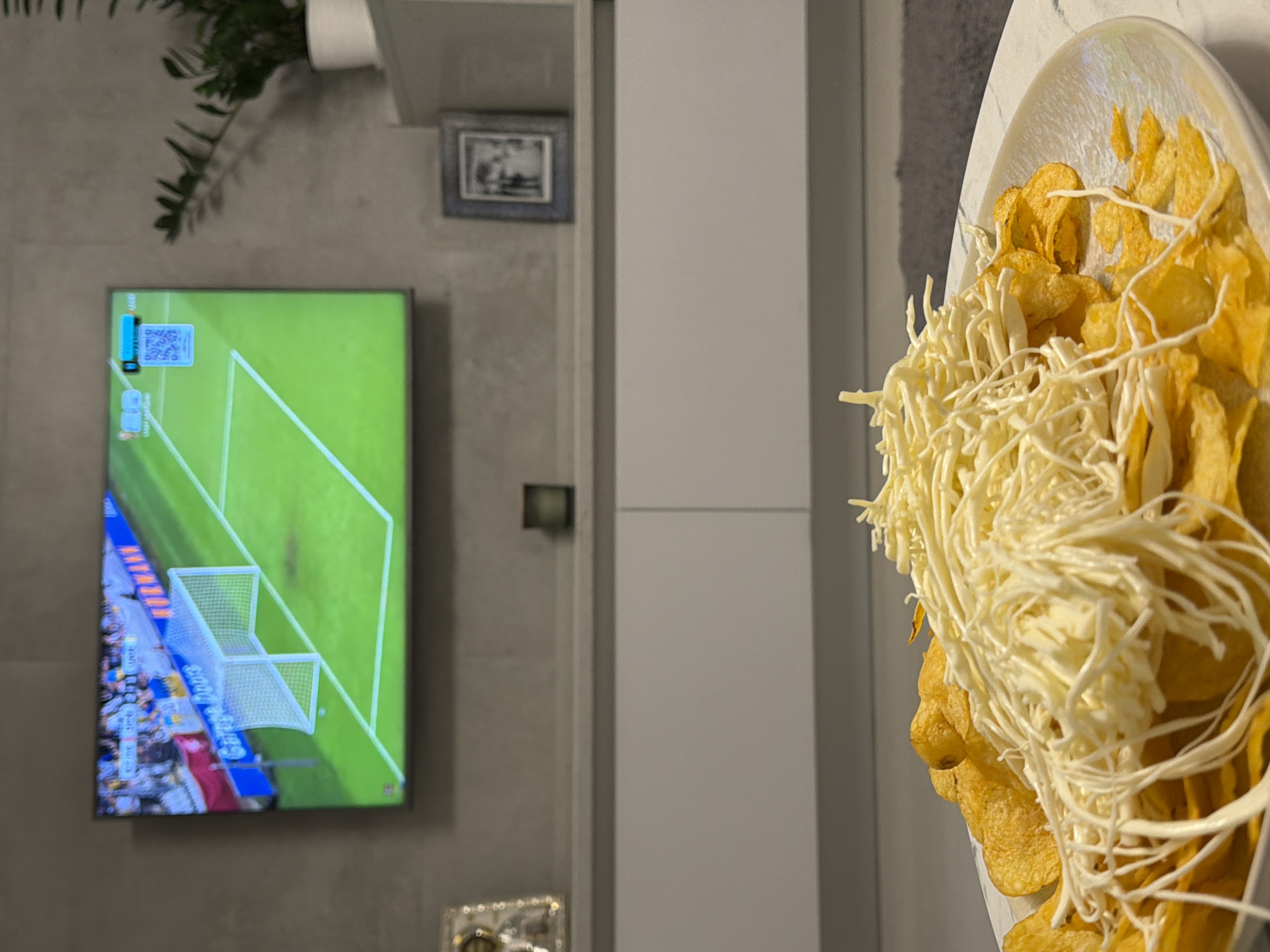 The scene is an interior view of a modern living space located in Sofiivska Borshchahivka, Ukraine. No individuals are present. In the immediate foreground, a round, white, iridescent plate is filled with golden potato chips, heavily covered by a large quantity of white, finely shredded cheese, consistent with string cheese. The plate rests on a light-colored, subtly patterned surface, likely a coffee table, with a dark grey rug partially visible to its right. The background features a textured, light grey wall. Mounted centrally on this wall is a large, flat-screen television displaying a soccer (football) match. The screen shows a green pitch with white markings and a white goal net. On-screen graphical elements include blurred text and numbers in the upper left, a blue rectangular graphic containing a QR code in the upper right, and spectator banners in the background of the pitch, one notably displaying "Garage Doors" alongside national colors, indicative of a live broadcast. Below the television, a long, white, minimalist media console extends across the wall. Objects on the console include a small, ornate, gold-framed icon on the left, a dark square object (possibly a candle) in the center, and a dark-framed photograph on the right. Further right, a potted plant with dark green leaves is positioned on an adjacent shelf. The environment suggests a domestic setting dedicated to leisure.