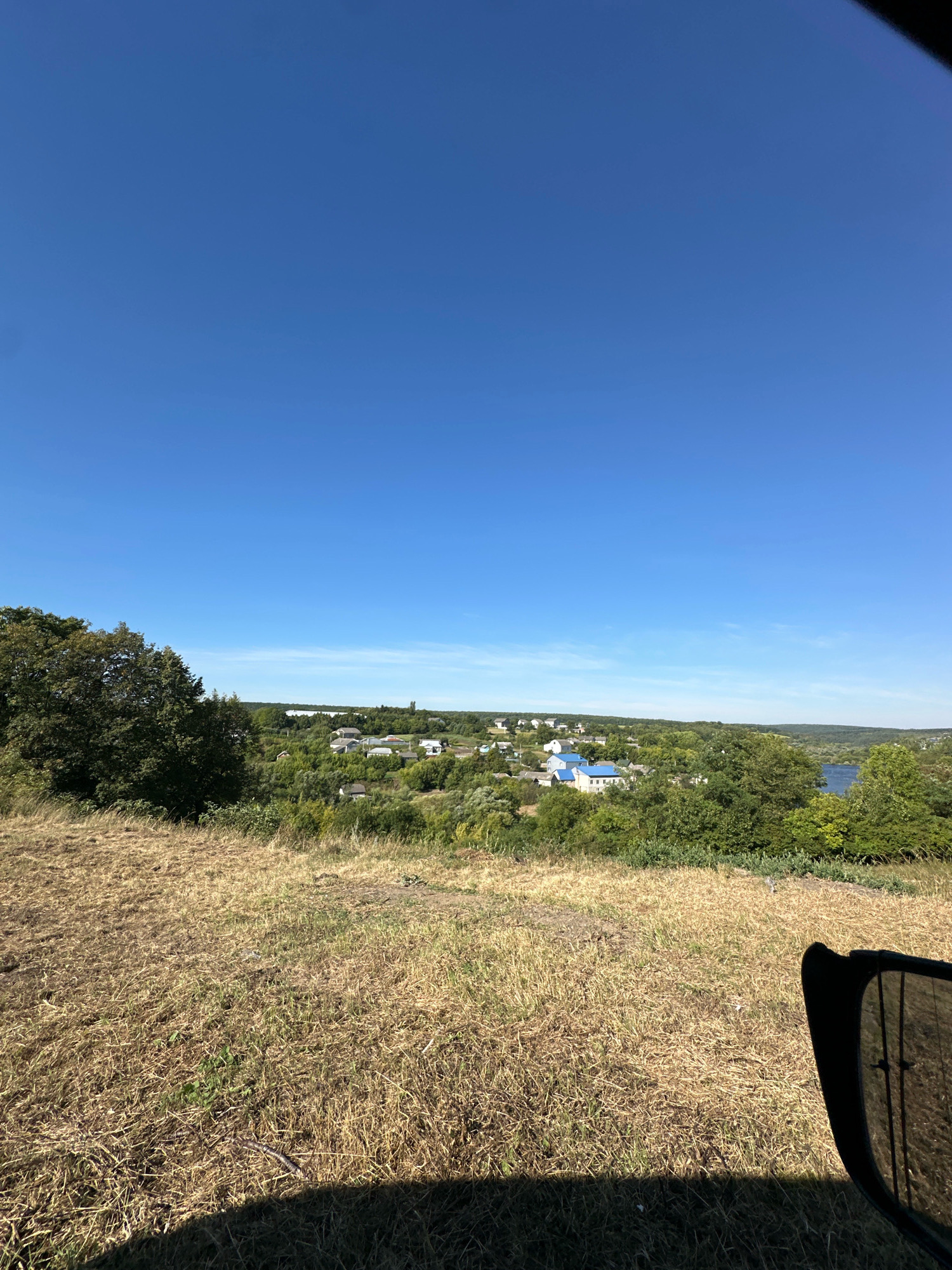 This is a wide-angle, eye-level outdoor photograph taken on a sunny day. The scene overlooks a rural village with houses and lush green trees. A body of water is visible in the distance, likely a river or lake. In the foreground, dry, golden grass covers a sloping hill. The sky is a clear, bright blue. Part of a dark, reflective object, possibly a car window or mirror, is visible in the lower right corner of the frame, casting a shadow on the grass. The location is identified as Makiv, Ukraine. There are no people or animals in the image, and no specific actions or interactions are depicted.