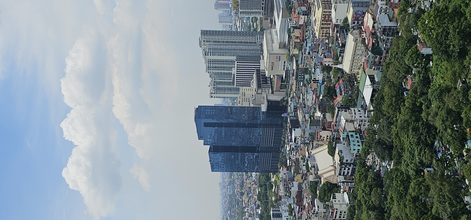The image depicts an urban landscape in Makati, Philippines, under a partially cloudy sky. In the foreground, dense green trees and foliage dominate the lower portion of the frame, suggesting a park or a heavily treed area within the city. Beyond this lush greenery, a densely packed residential and commercial area unfolds, characterized by a mix of low-rise buildings with colorful roofs and multi-story structures.
Dominating the midground are several modern skyscrapers. The most prominent is a dark blue, glass-faced building with a distinctive tiered design, likely an office tower. To its right, a cluster of lighter-colored, taller residential buildings with a repeating balcony pattern stands out. Further in the background, more high-rise buildings and the general urban sprawl of Makati are visible, receding into a hazy distance.
Among the lower buildings, a white, angular structure with a prominent roofline, possibly a church or a community hall, is visible. Other buildings exhibit a variety of architectural styles and colors, indicative of a dynamic and developed urban environment. The overall scene captures the layered nature of Makati, where natural elements coexist with a dense, modern cityscape. There are no people or active events visible in the image.
