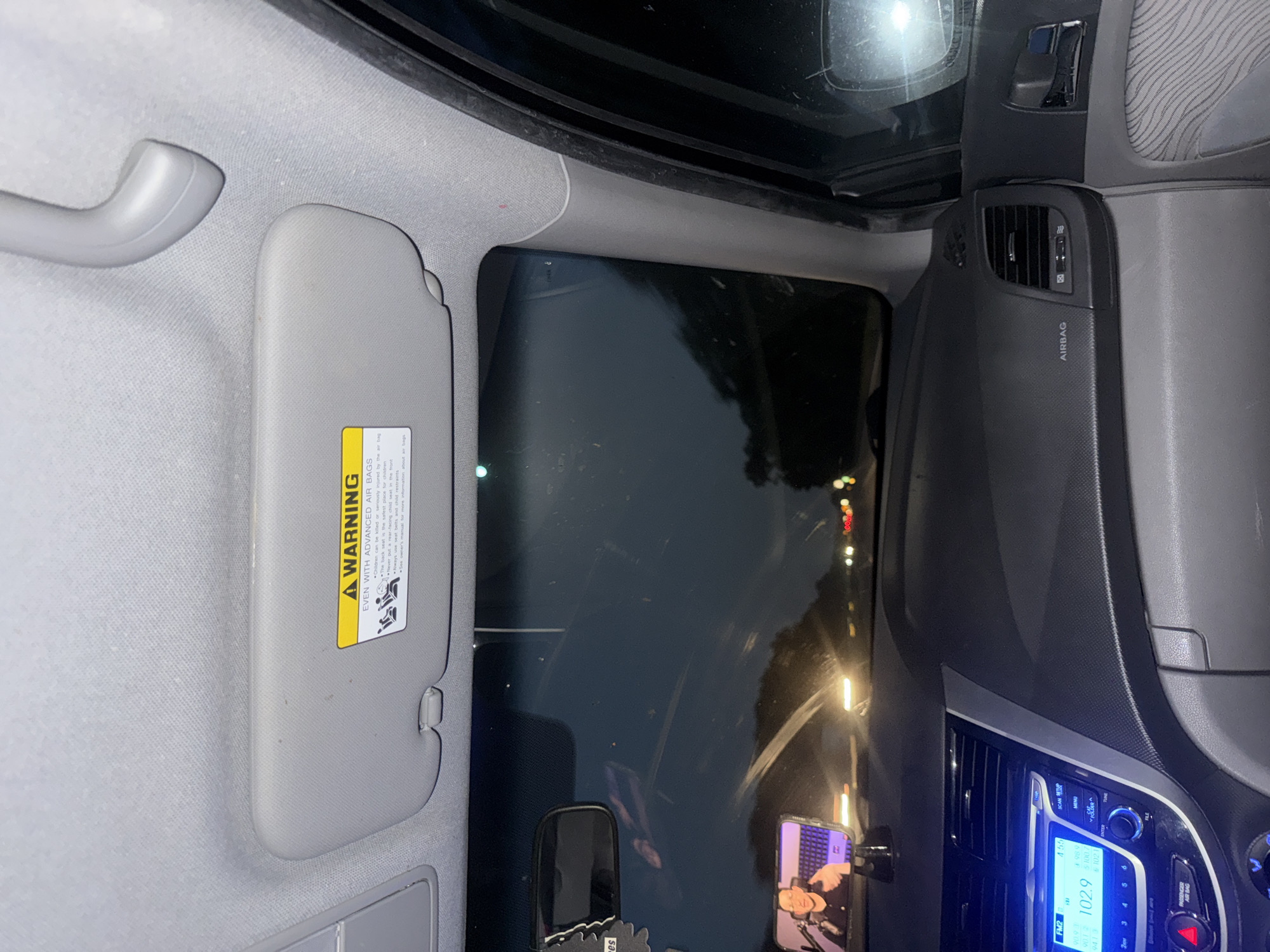 The image shows the interior of a car at night. The view is from the passenger seat, looking forward. The dashboard, center console, and part of the driver's side door are visible. A smartphone is mounted on the dashboard, displaying a video of a person. The windshield reflects the dark sky and the headlights of other vehicles on the road. The car appears to be moving. A warning sticker is visible on the passenger side visor. The car is located in Manheim Township, United States of America.
