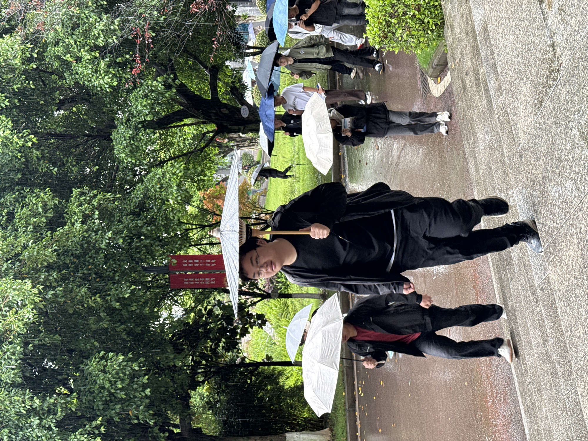 On a damp, overcast day in Jiangjin, China, a group of people, mostly young adults, are walking outdoors under umbrellas. The scene appears to be on a path near a park or wooded area, as evidenced by the lush green trees on the left side of the frame. Some of the people are holding white, decorative umbrellas, while others have more traditional rain umbrellas, some blue or black. The path is paved and appears wet from recent rain. The individuals are dressed in a variety of casual clothing, some in black, while others wear lighter colors. In the background, a set of stairs leads upwards, and another group of people can be seen walking away. A red sign with Chinese characters can be seen among the trees.