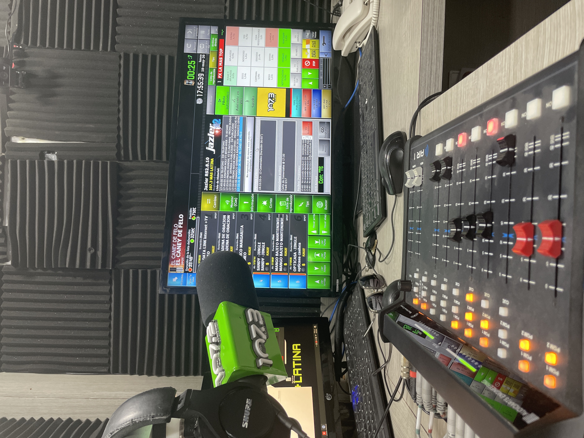 The image shows a radio broadcasting studio in Porlamar, Venezuela. A "EZQI LATINA" branded microphone is positioned in front of a computer monitor displaying Jazler radio automation software with a playlist, including tracks like "EL CANEY DE FELO". A mixing console labeled "AIR 1 AUDIOARTS ENGINEERING" is visible, with multiple faders and illuminated buttons. A pair of SHURE headphones rests near the microphone. A second computer monitor displays program information and automation controls. Other objects visible in the studio include a keyboard, computer mouse, telephone, and a pair of sunglasses. The walls are covered in soundproofing material.