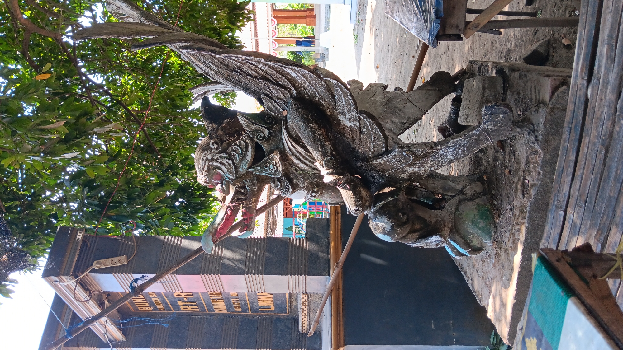 In Kelurahan, Indonesia, a weathered, grey stone statue of a mythical creature resembling a garuda stands prominently in an outdoor setting. The statue is positioned on a concrete pedestal, which is slightly elevated from the dirt ground. The creature has large wings, a bird-like head with an open beak revealing red and white details, and a powerful, muscular body. It appears to be crushing a smaller, rounded figure beneath its claw.
Behind the statue, a dark grey pillar with horizontal indentations and gold lettering is visible. The lettering appears to be names or designations, with "RT-R" and "KARANGTAR" being partially legible. A wooden pole runs horizontally across the scene in front of this pillar and the statue. Lush green foliage from a tree with dappled sunlight filters through the branches, casting shadows on the statue and its surroundings.
In the background, a light-colored building with red and white bunting is discernible, along with glimpses of colorful playground equipment, suggesting the area might be near a public space or school. The ground is a mix of dirt and sand, with some scattered debris. In the foreground, a portion of a bamboo seating arrangement is visible, covered with a patterned mat, indicating a place for rest or gathering. The overall impression is a cultural or religious landmark in a village or semi-urban Indonesian environment.
